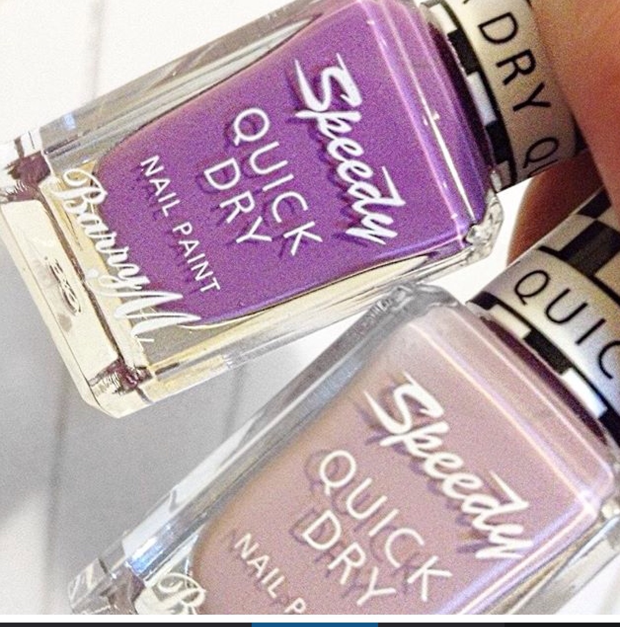 Two Barry M Nail polishes being held against a white background. One is in a lilac shade whilst the other is a darker shade of purple