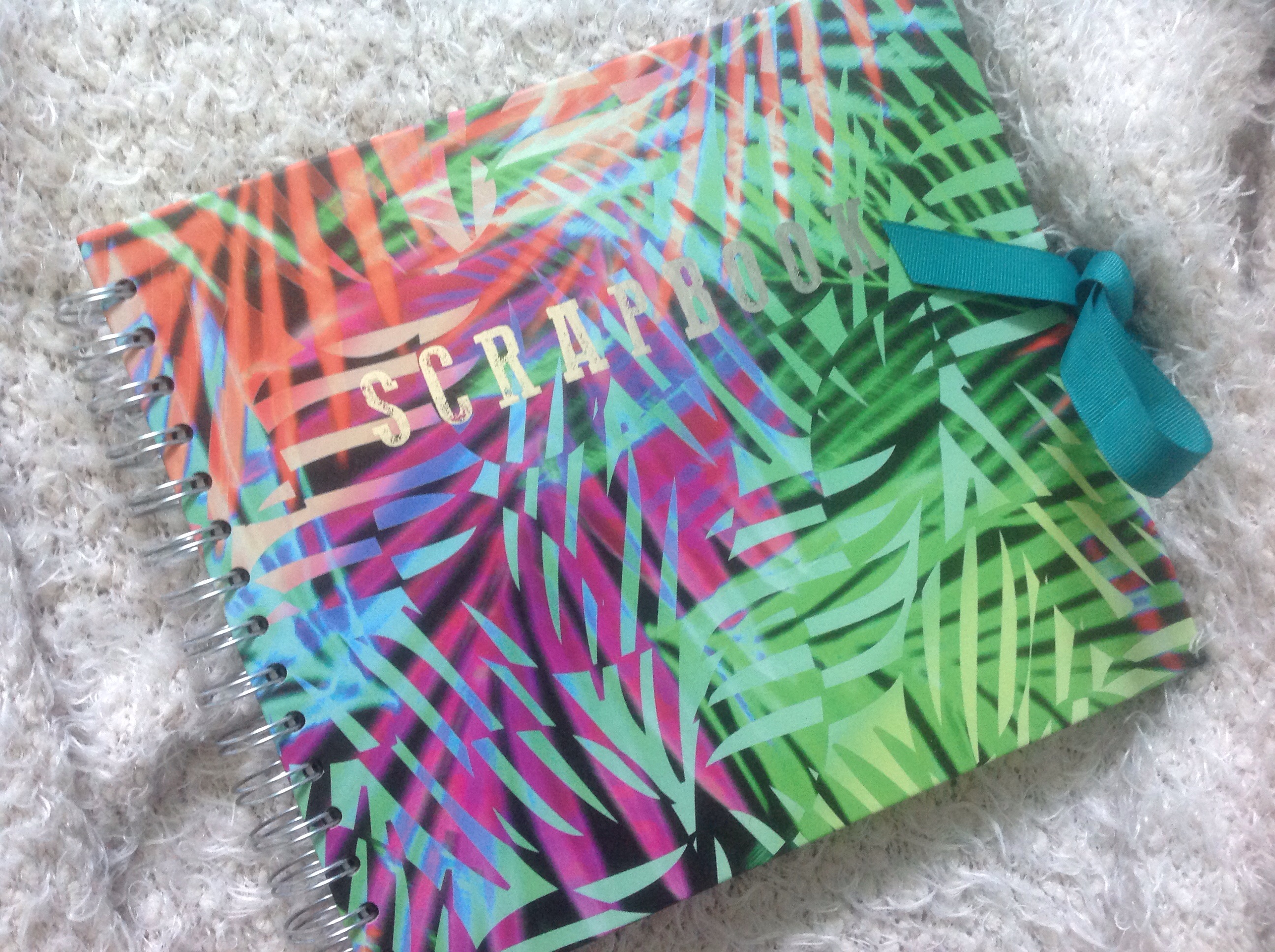 A brightly coloured scrapbook which has the pattern of exotic tree leaves in pink, orange, light blue and green. The word Scrapbook is scribed in silver block letters in the centre