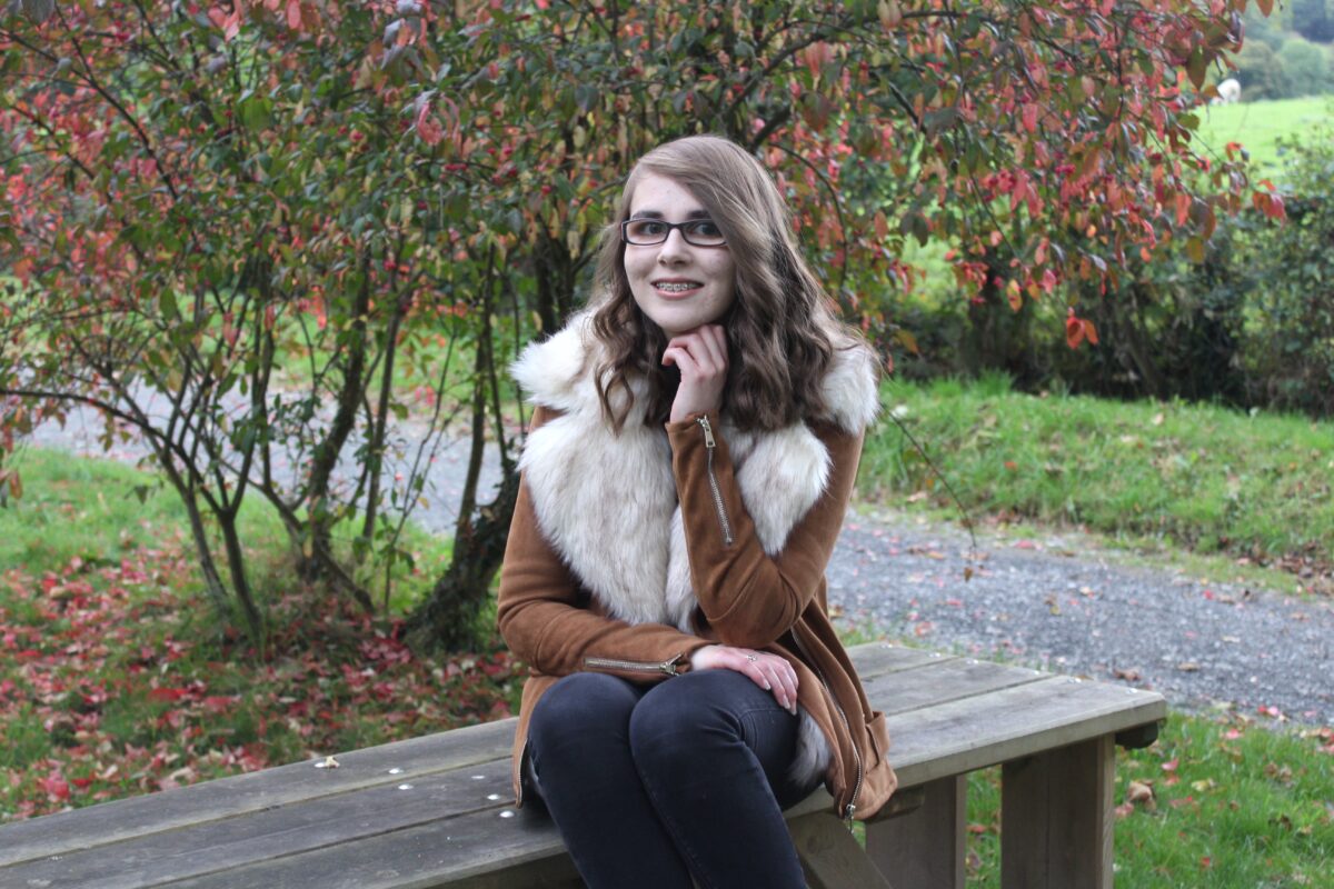 Elin sitting on a bench wearing a tan faux suede aviator jacket with cream fur lining