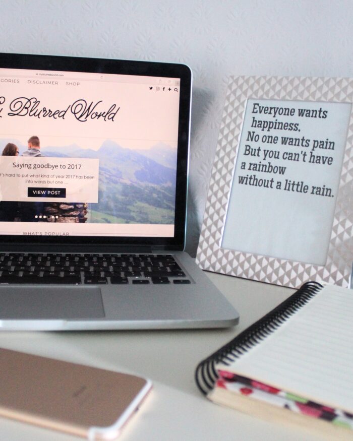 A photo of my blog on a macbook, there is a quote in a frame next to it that says everyone wants happiness, no one wants pain but you can't have a rainbow without a little rain