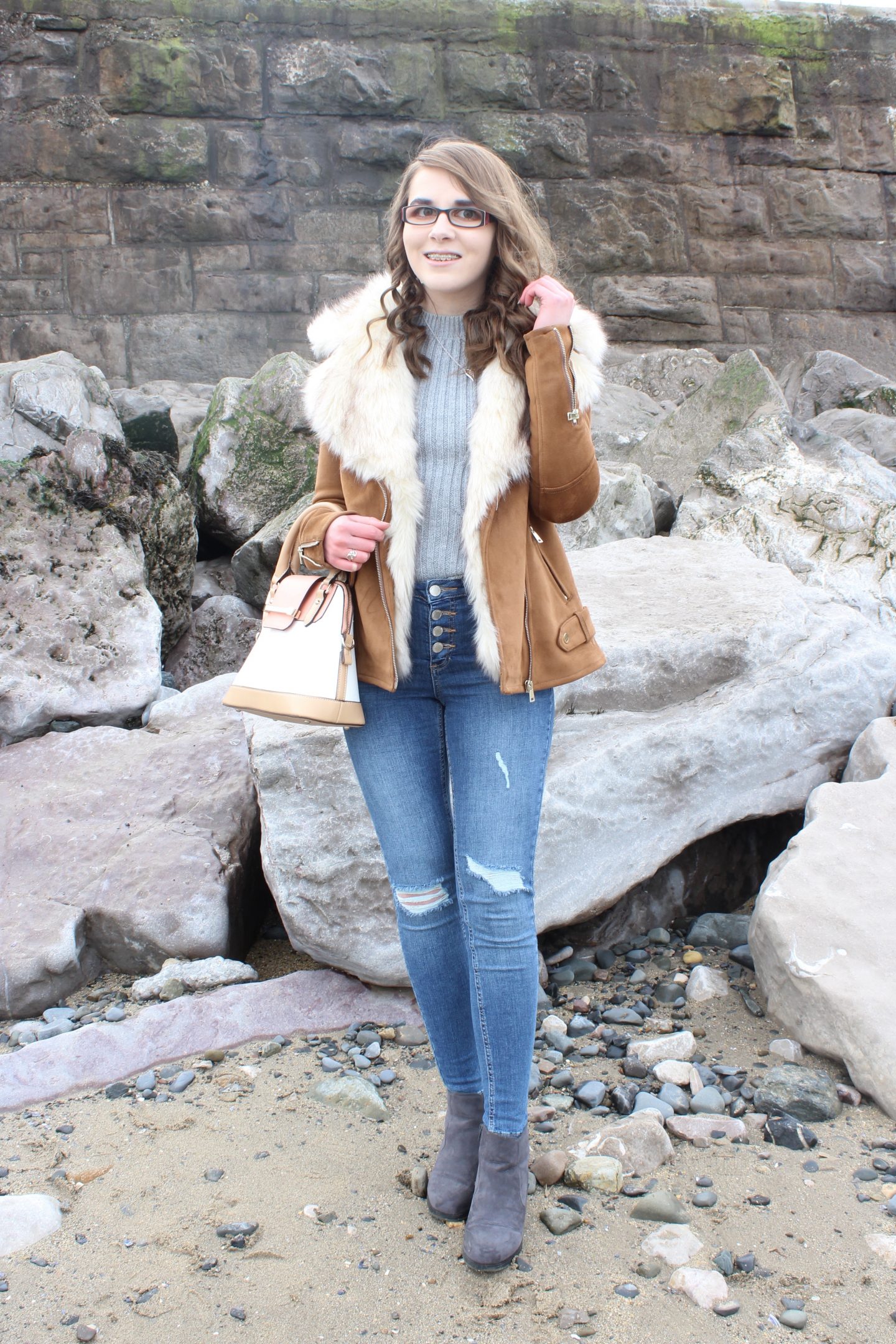 A photo of Elin wearing a tan faux suede and faux fur trim jacket, a grey knitted jumper, blue ripped jeans, grey ankle boots and a cream, peach and tan handbag