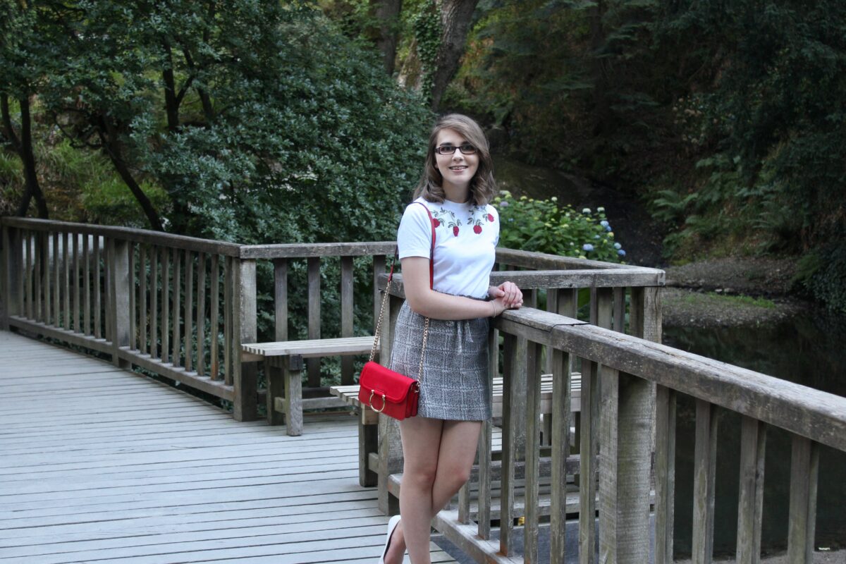 A photo of Elin leaning on a bridge, wearing a white t-shirt with red embroidered flowers on the neckline, a paperbag skirt and small red handbag with gold ring detail and white slip on shoes