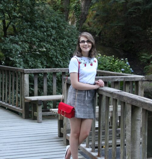 A photo of Elin leaning on a bridge, wearing a white t-shirt with red embroidered flowers on the neckline, a paperbag skirt and small red handbag with gold ring detail and white slip on shoes