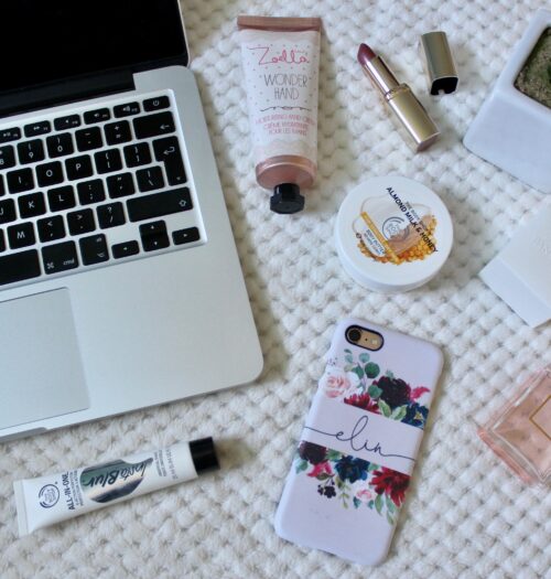 A photo of a macbook keyboard with beauty products around it and my phone which has a pink case and my name on it