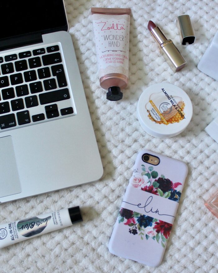 A photo of a macbook keyboard with beauty products around it and my phone which has a pink case and my name on it