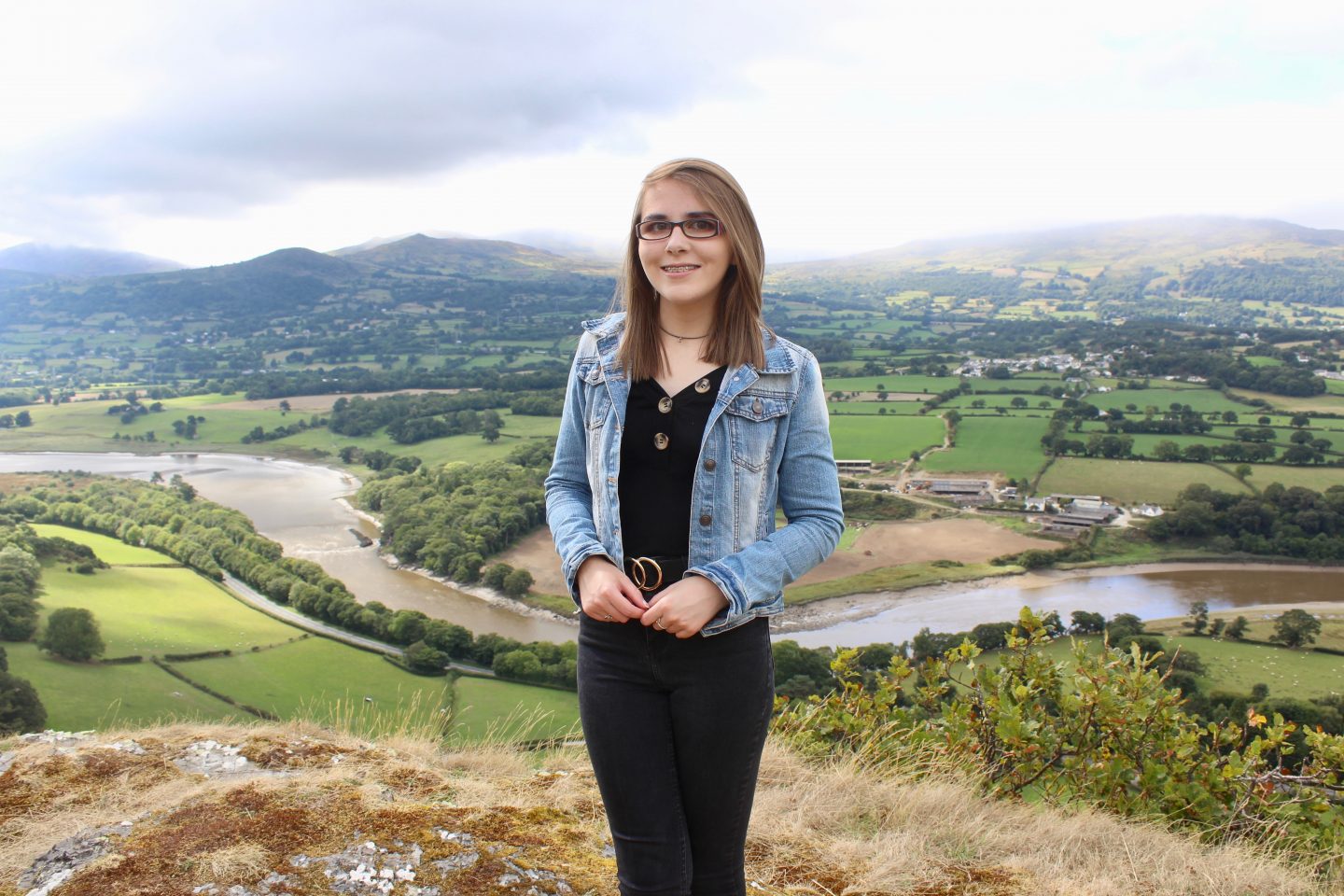 A photo of Elin wearing black top, skinny jeans and a denim jacket. There is a river and mountains in the background