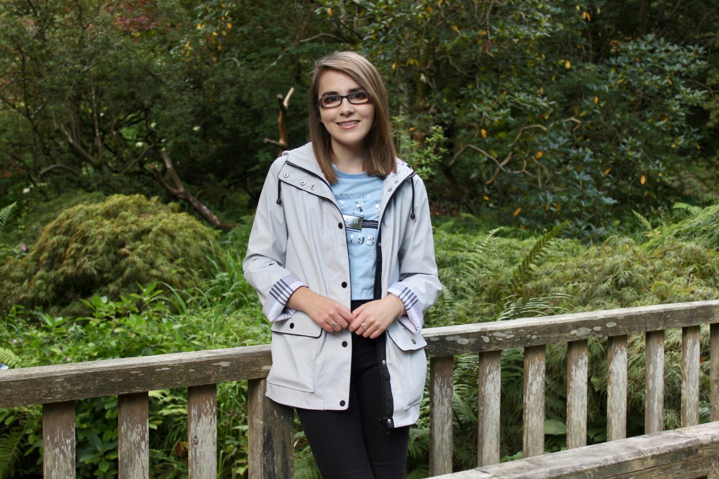 A photo of Elin leaning on a bridge wearing a light grey raincoat with check detailing on the end of the sleeves, a light blue top with silver writing on it and black skinny jeans