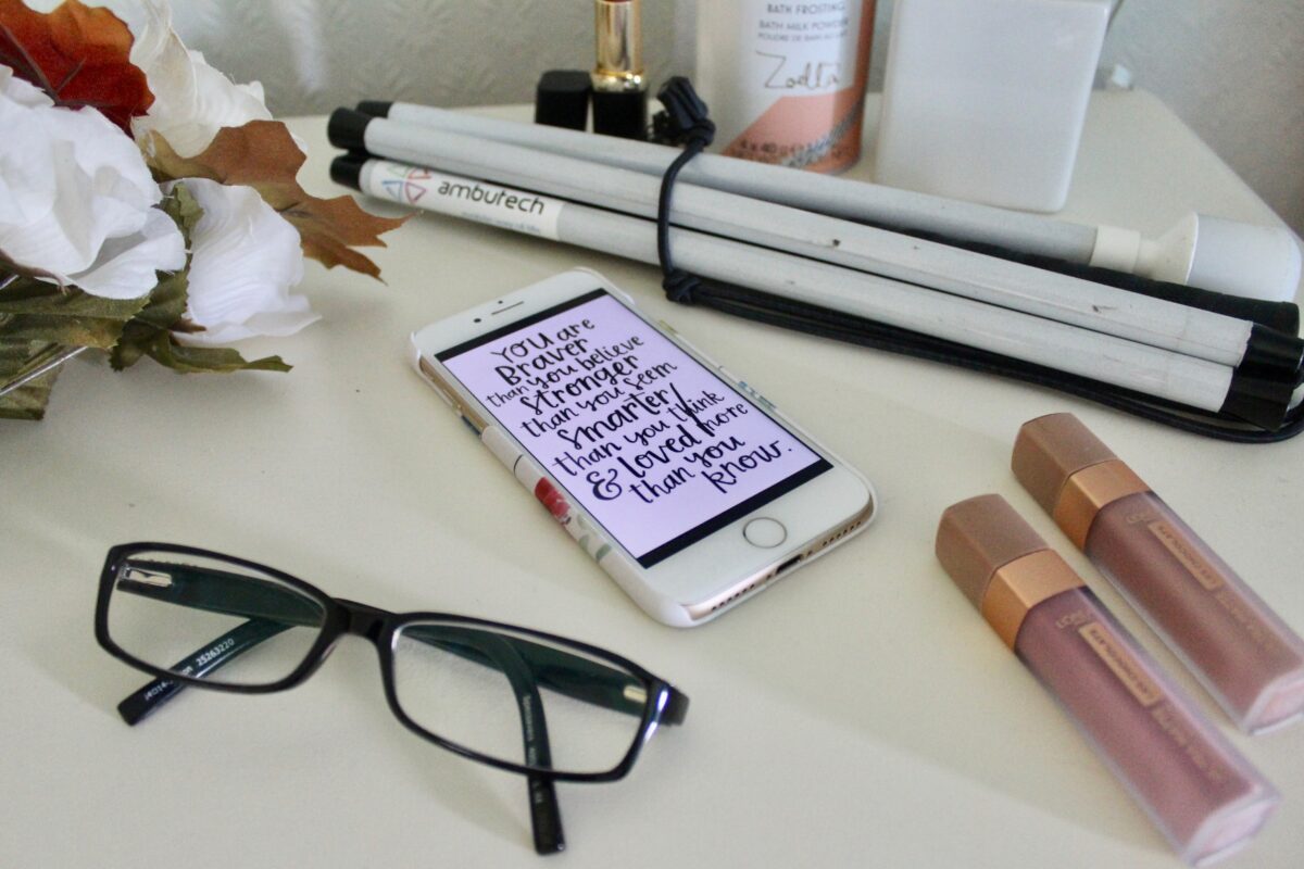 A photo of a black pair of glasses with my phone beside it which has a quote which says 'you're braver than you believe, stronger than you seem, smarter than you think and loved more than you know', my cane is behind my phone and there are a few beauty products dotted around
