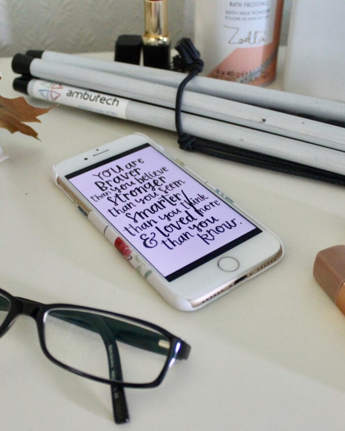 A photo of a black pair of glasses with my phone beside it which has a quote which says 'you're braver than you believe, stronger than you seem, smarter than you think and loved more than you know', my cane is behind my phone and there are a few beauty products dotted around