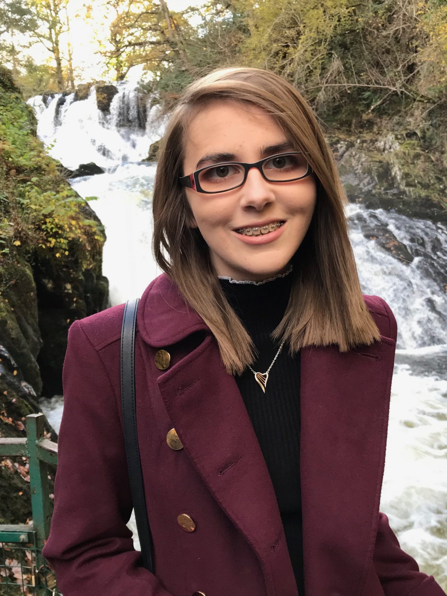 A portrait photo of Elin wearing a high neck black top with frill detailing on the collar, a burgundy coat and harp shaped necklace