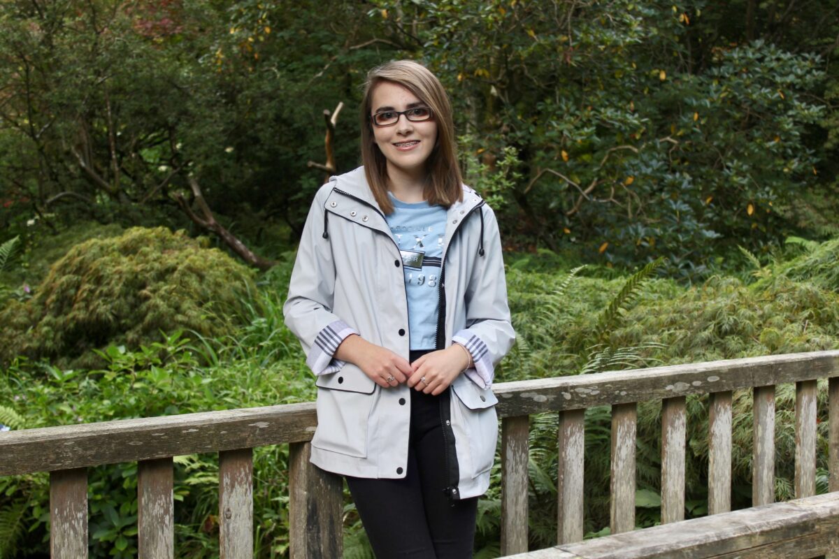 A photo of Elin in a grey raincoat standing on a wooden bridge