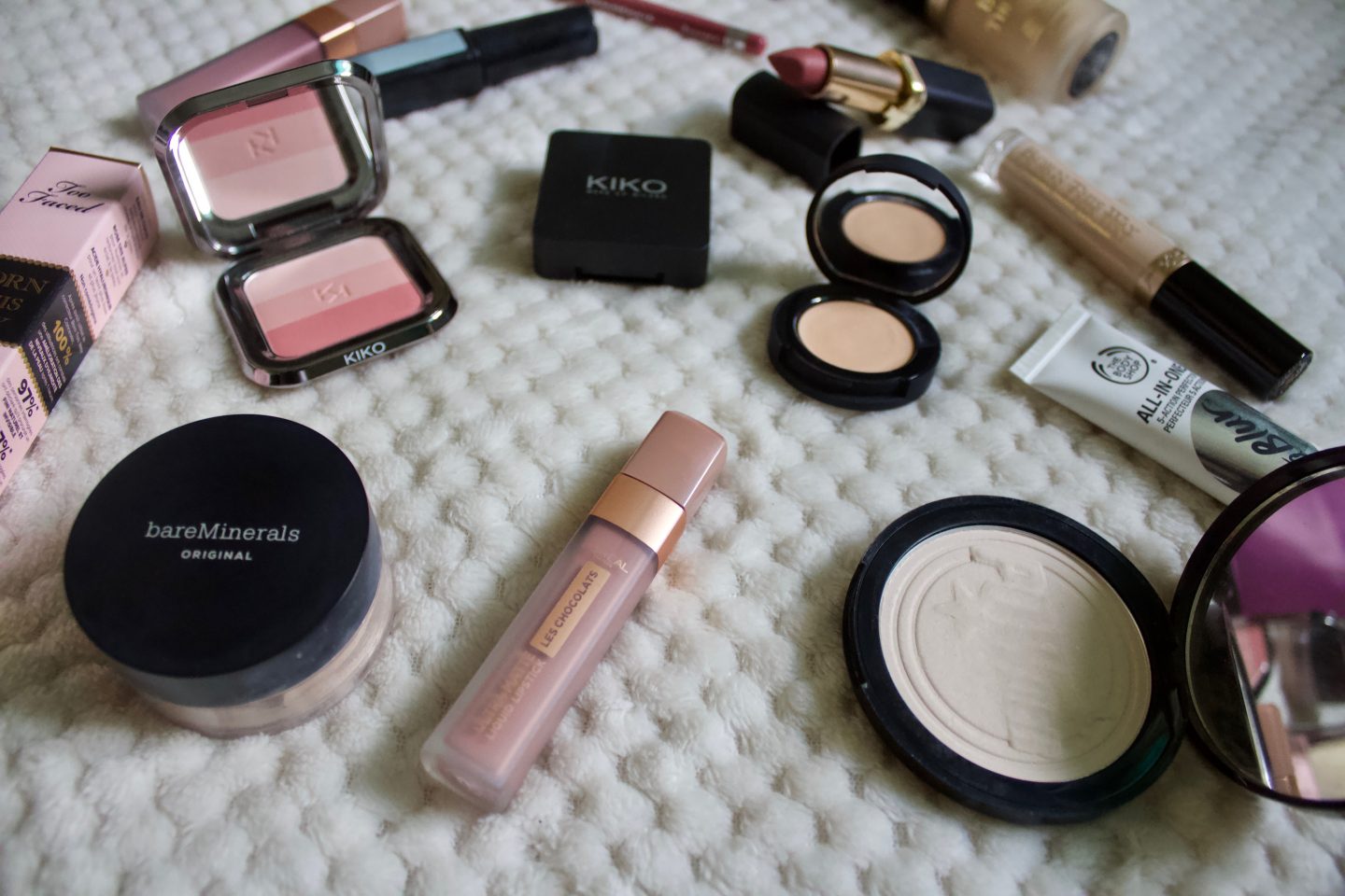 Different makeup products including a l'oreal liquid lipstick, S&G powder, bare minerals foundation and kiko single eyeshadow. They're placed on a textured cream throw