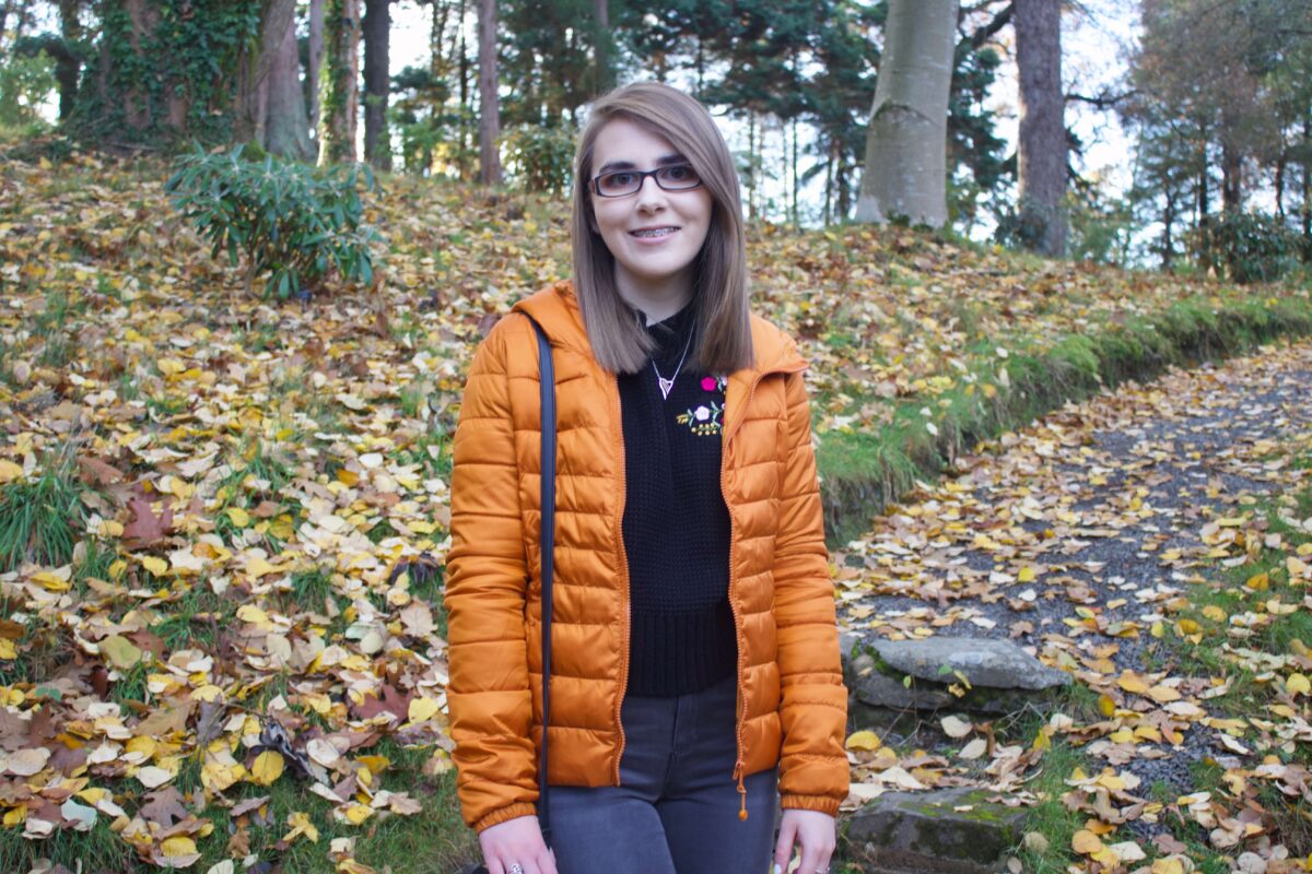A photo of Elin wearing a black knitted jumper and orange/marmelade coloured quilted jacket, she is standing on some steps in a woodland and there are colourful leaves on the floor around her.