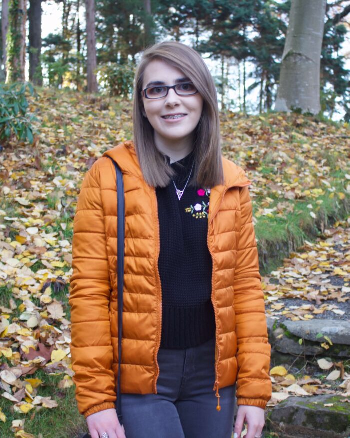 A photo of Elin wearing a black knitted jumper and orange/marmelade coloured quilted jacket, she is standing on some steps in a woodland and there are colourful leaves on the floor around her.