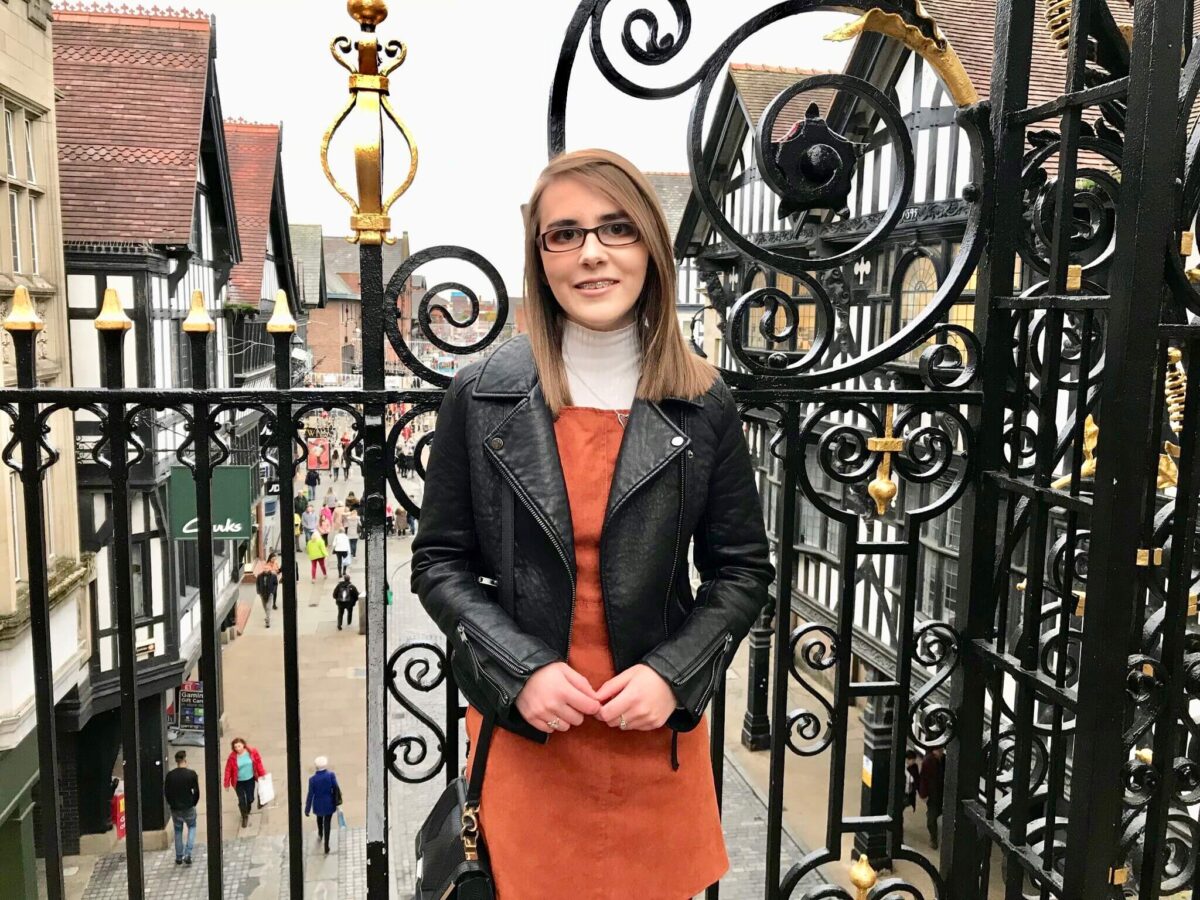 A photo of Elin in a burnt orange pinafore and black leather jacket. She is standing in front of pretty black railings with a street in the background