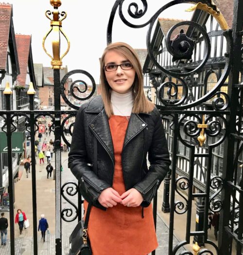A photo of Elin in a burnt orange pinafore and black leather jacket. She is standing in front of pretty black railings with a street in the background