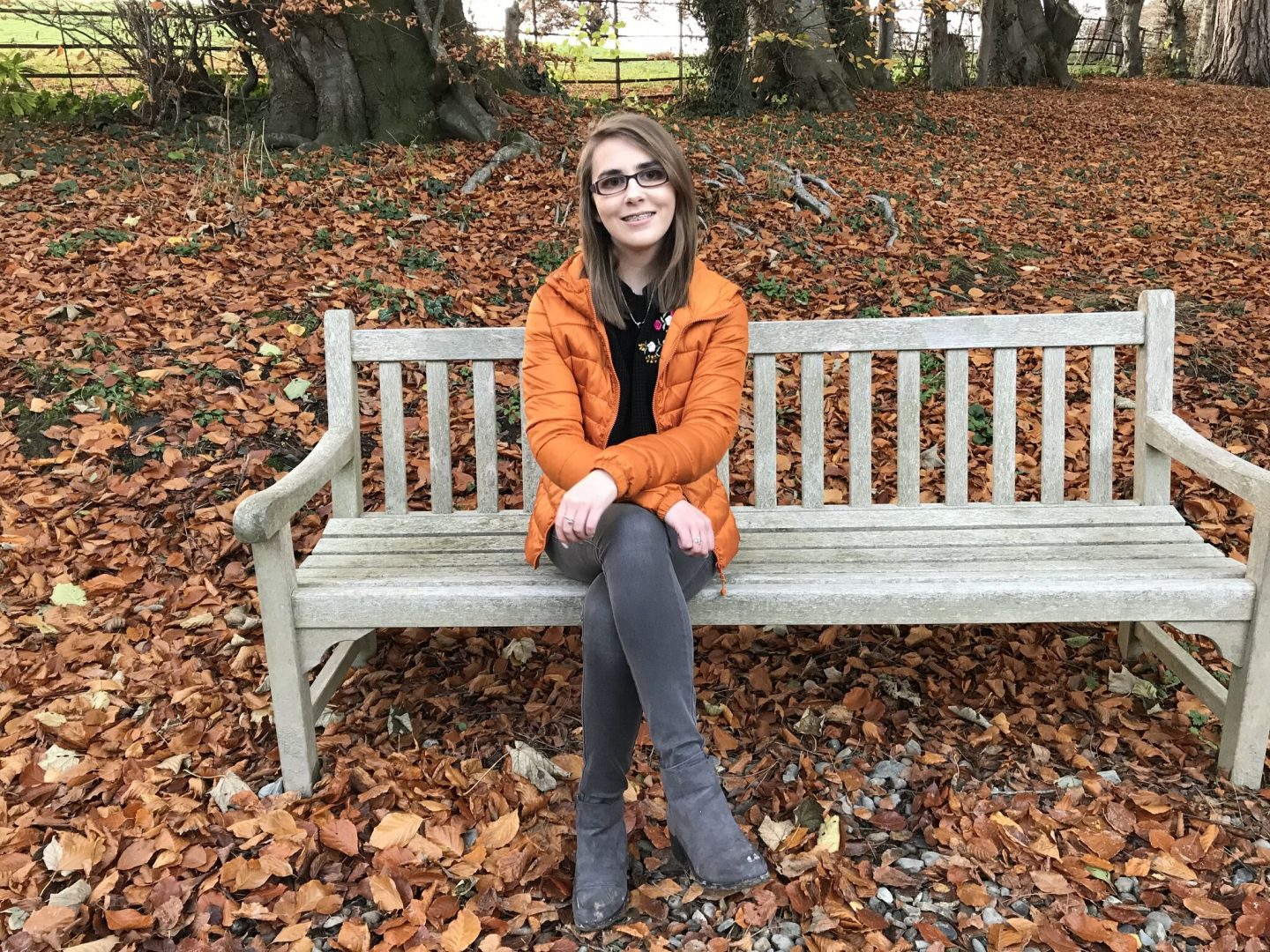 Elin sitting on a wooden bench which has an array of colourful leaves scattered around it