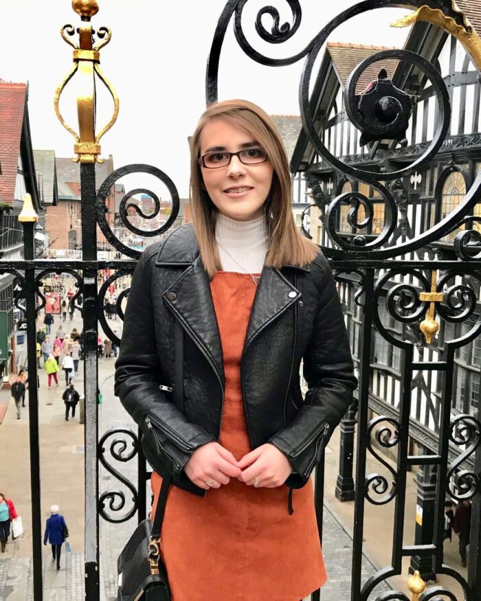 A photo of Elin in a burnt orange pinafore and black leather jacket. She is standing in front of pretty black railings with a street in the background