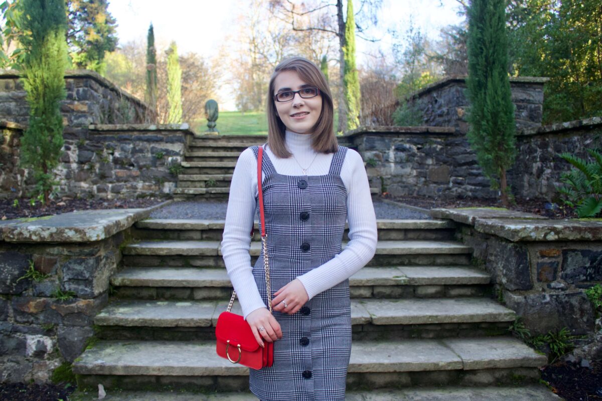A photo of Elin wearing a grey check button up pinafore, a high neck white long sleeve top and red handbag which has a gold ring on the front.