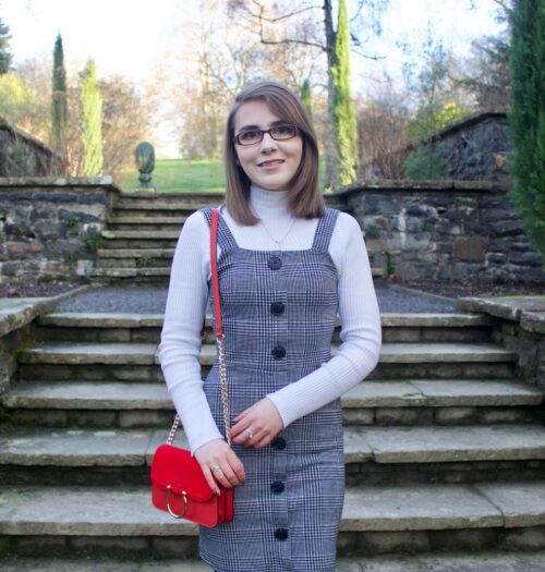 A photo of Elin wearing a grey check button up pinafore, a high neck white long sleeve top and red handbag which has a gold ring on the front.