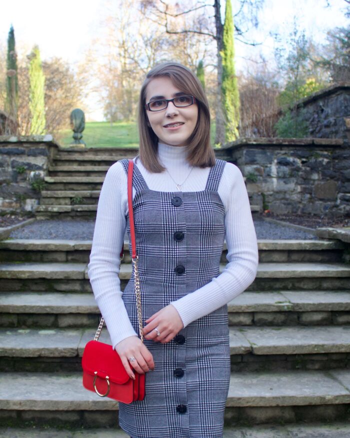 A photo of Elin wearing a grey check button up pinafore, a high neck white long sleeve top and red handbag which has a gold ring on the front.