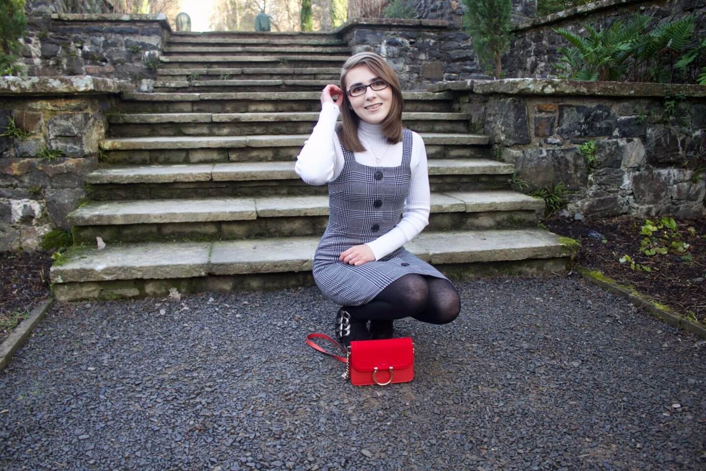A photo of Elin crouching down in front of some light steps, her red handbag is place upright on the floor in front of her