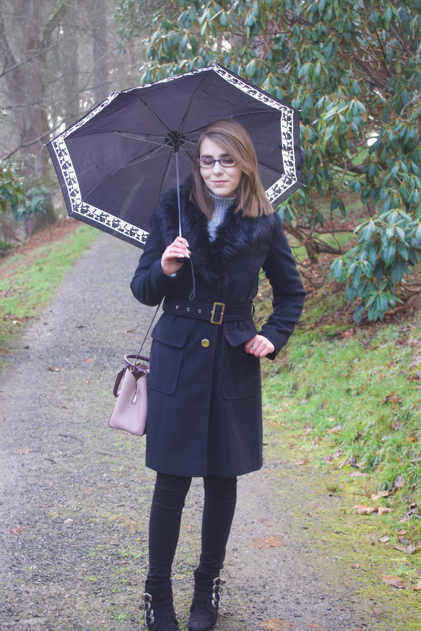 A photo of Elin looking down, holding an umberella. She is wearing a black longline coat which has a faux fur collar, black skinny jeans, a grey high neck knitted jumper and a Michael Kors bag
