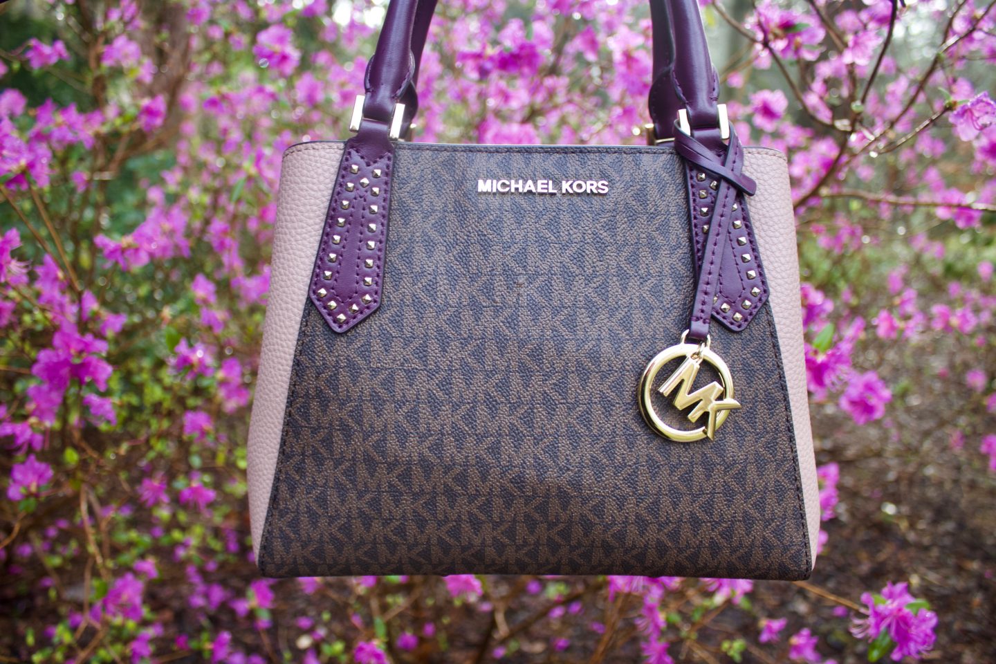 A photo of a brown Michael Kors bag wich has light pink sides and purple handles. There are pink flowers in the background