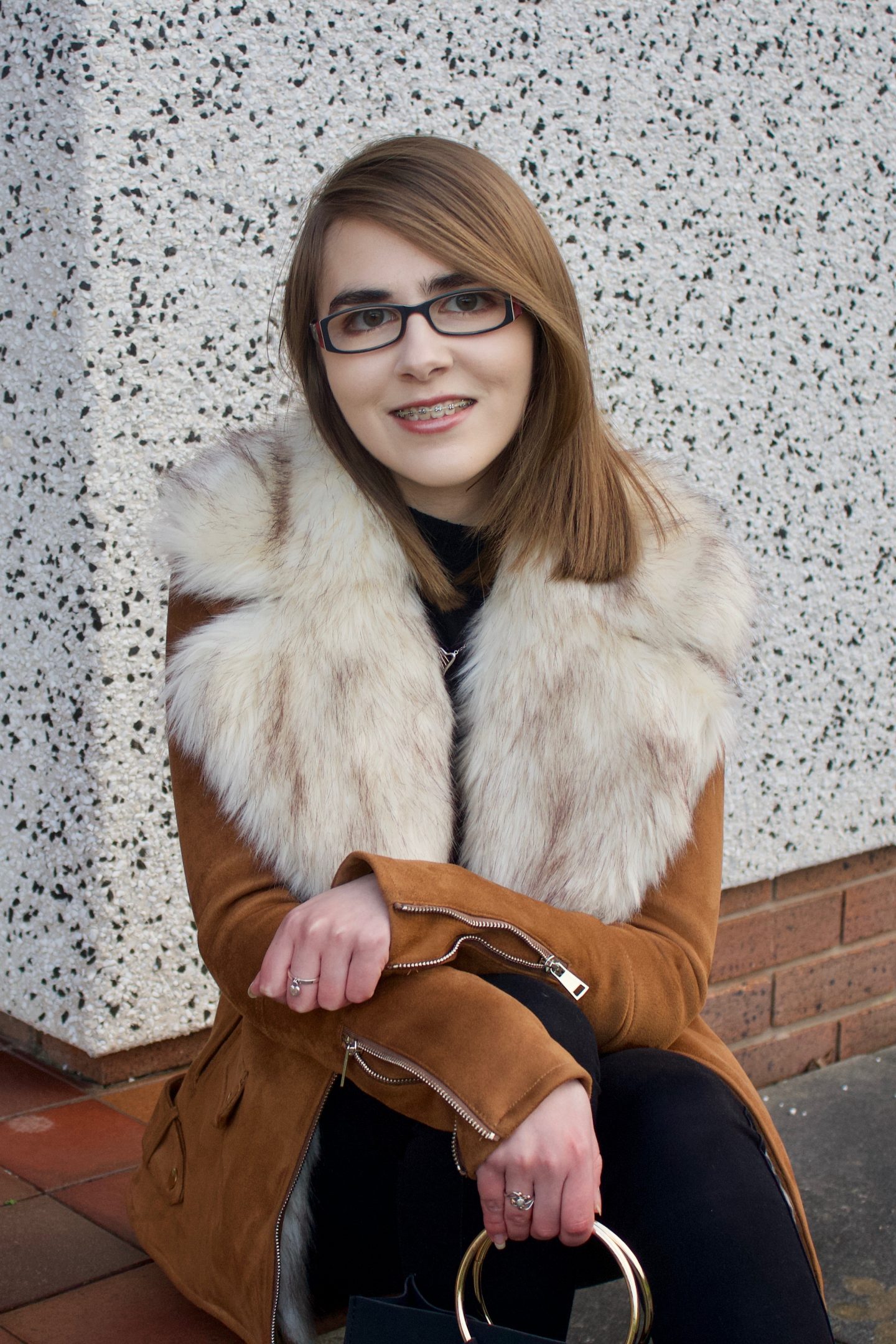 A photo of Elin sat on a step wearing a brown suede jacket which has a big faux fur collar