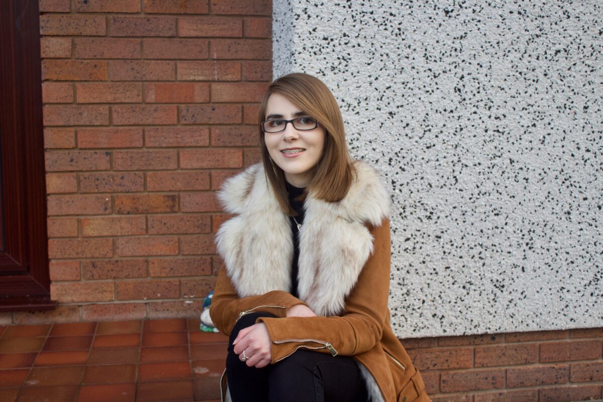 A photo of Elin sat on a step wearing a brown suede jacket which has a big faux fur collar, there is a red brick wall in the background in contrast to a white wall beside it