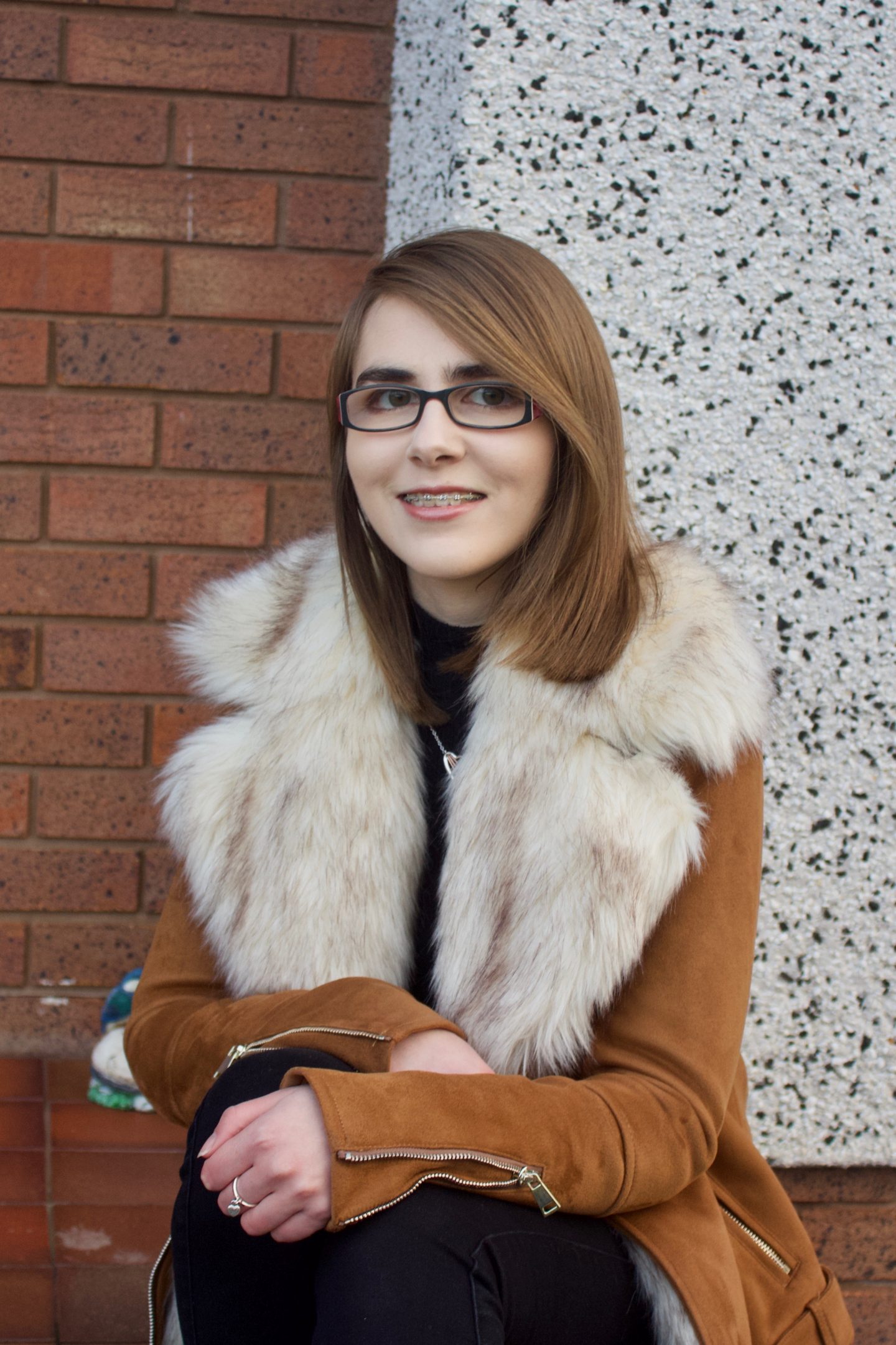 A photo of Elin sat on a step wearing a brown suede jacket which has a big faux fur collar