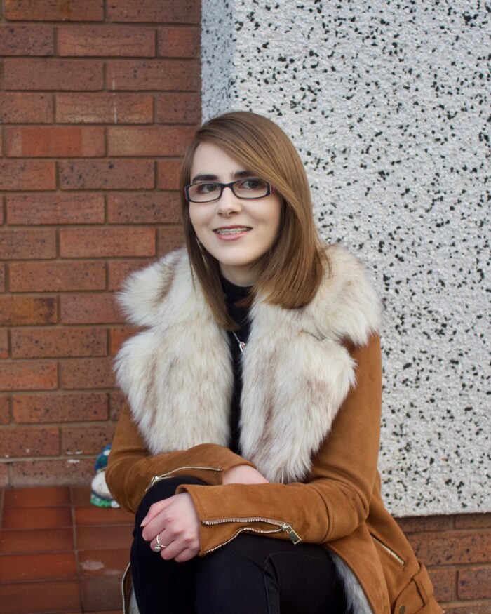 A photo of Elin sat on a step wearing a brown suede jacket which has a big faux fur collar, there is a red brick wall in the background in contrast to a white wall beside it