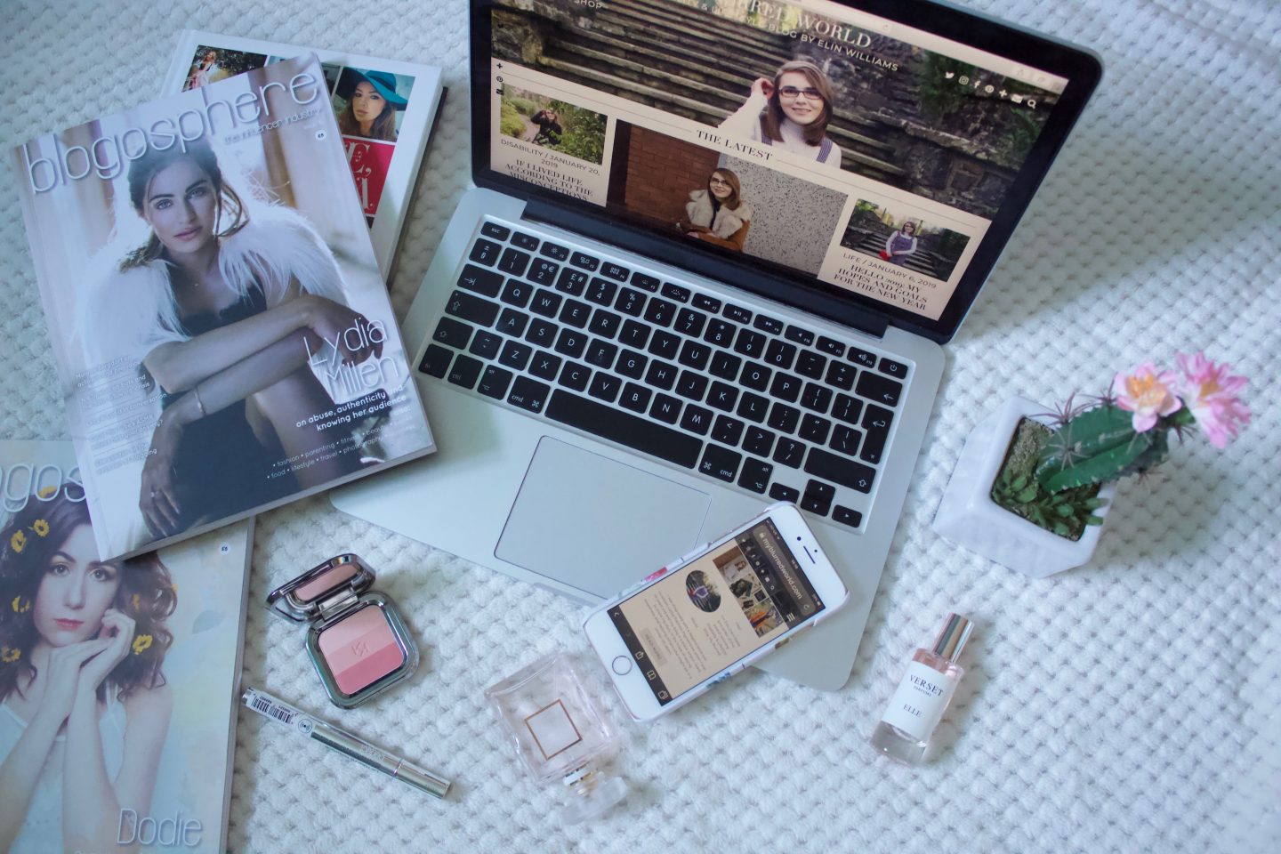 A photo of my blog on a macbook, there are a couple of issues of Blogosphere Magazine beside it, some makeup products and a fake cactus