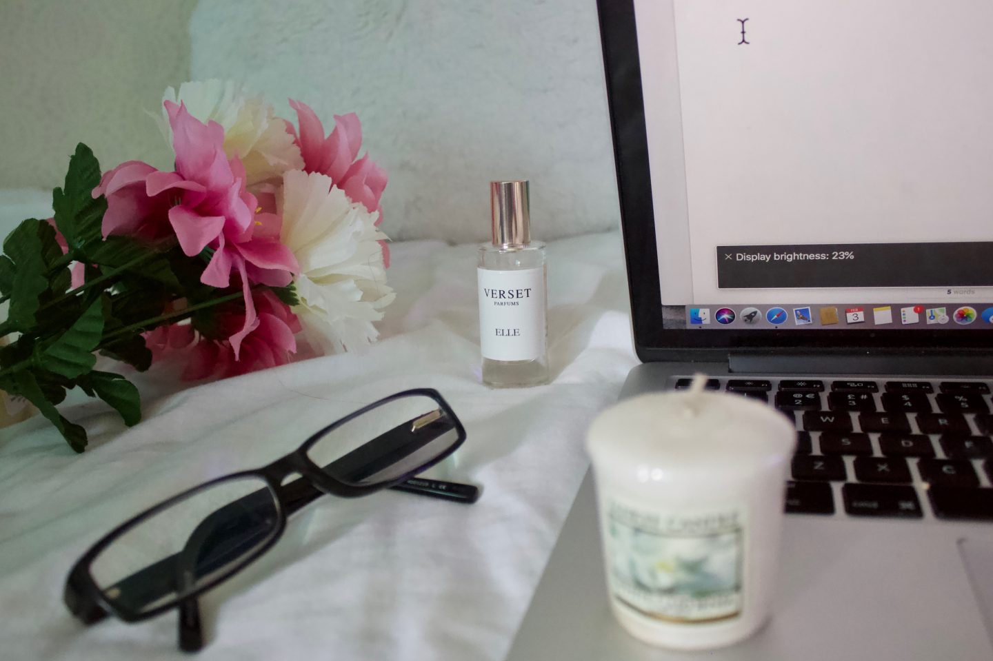 A photo of a pair of black glasses, there are fake flowers and a perfume in the background and a small candle which is upright on my macbook