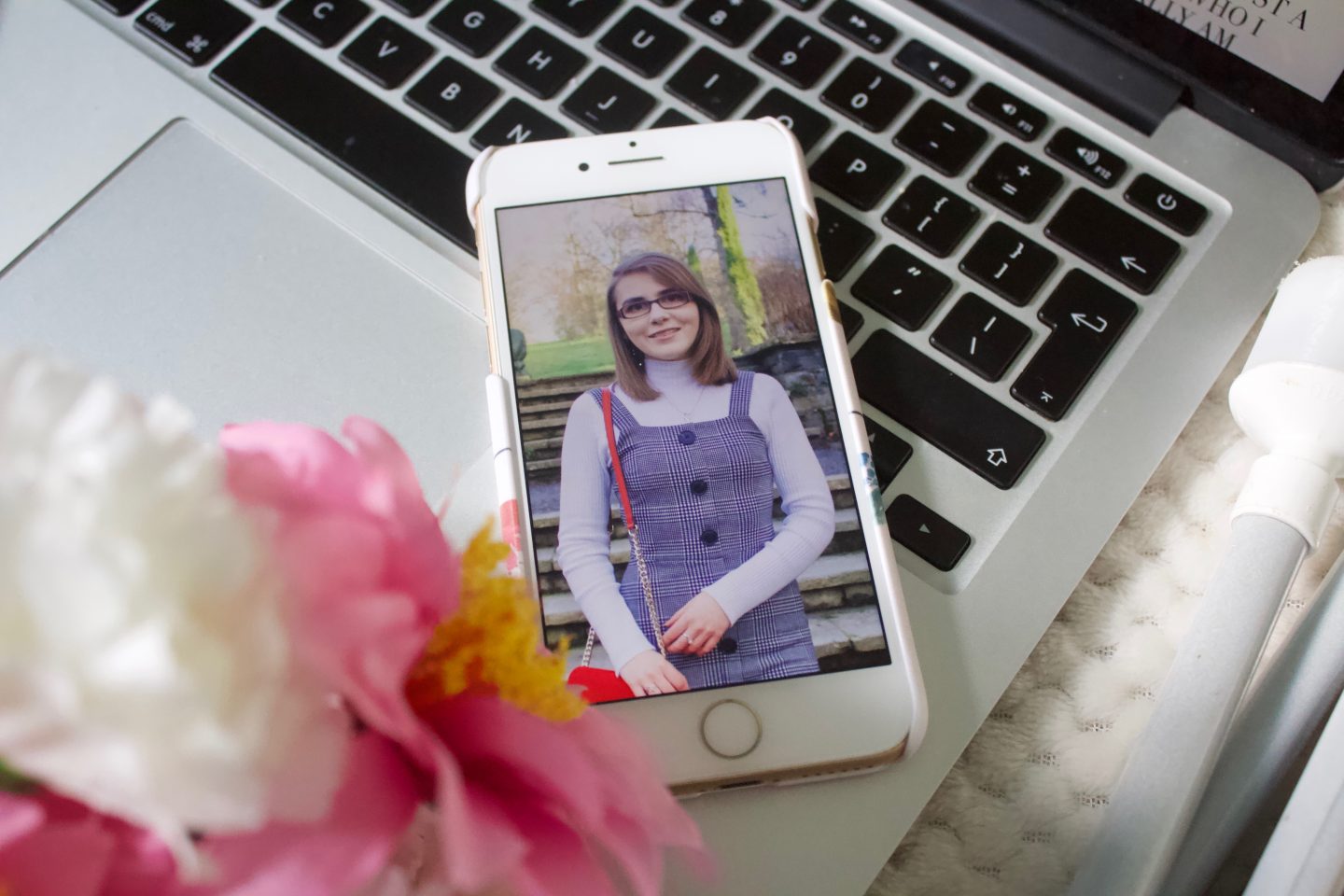 A photo of me in a check button up pinafore shows on my phone, there are some fake pink and white flowers in the left corner of the photo and my phone is placed on my macbook keyboard