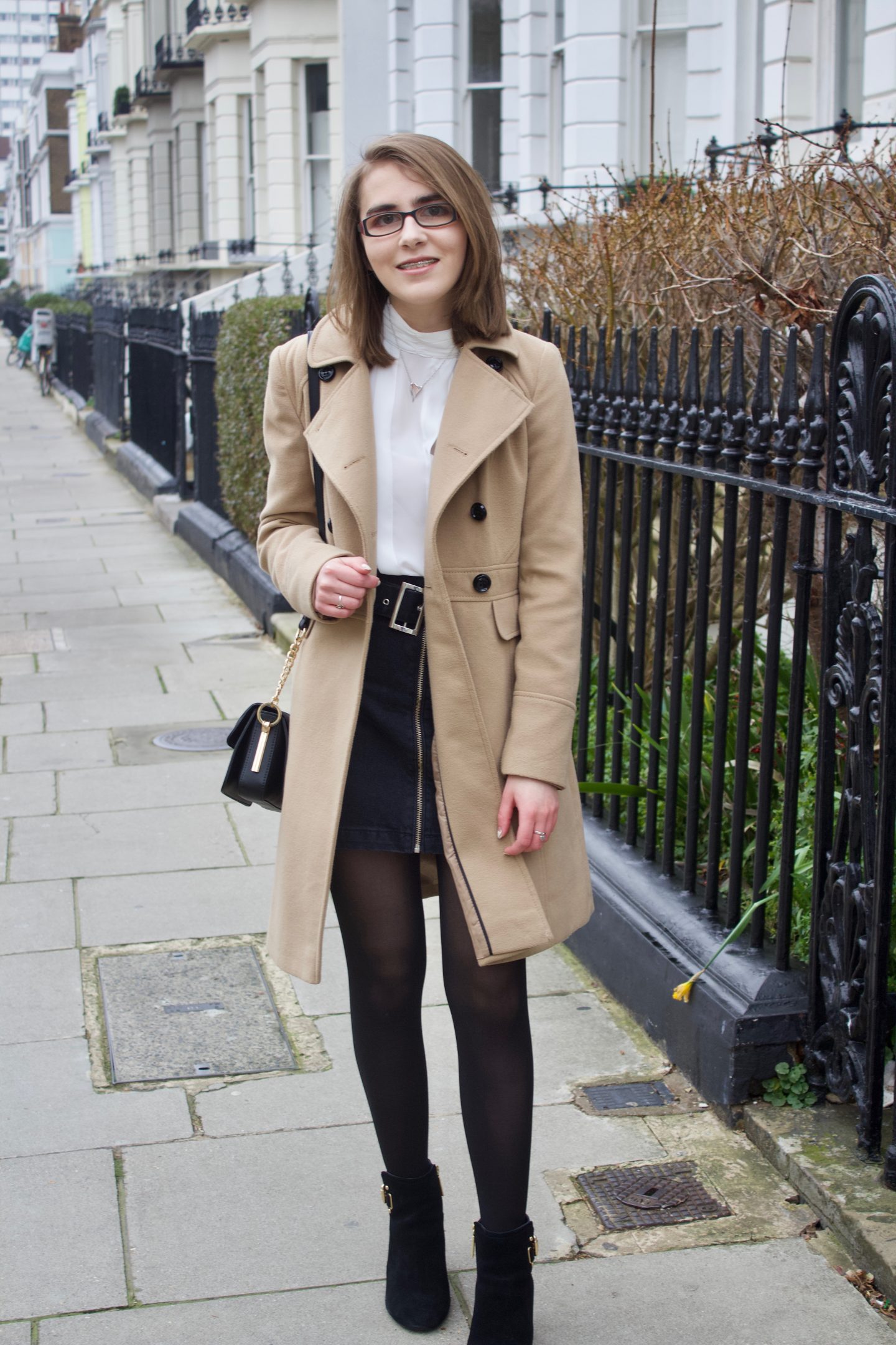 Elin, she is wearing a double breasted camel coat, a white pleated high neck blouse, a black denim skirt and black heeled ankle boots