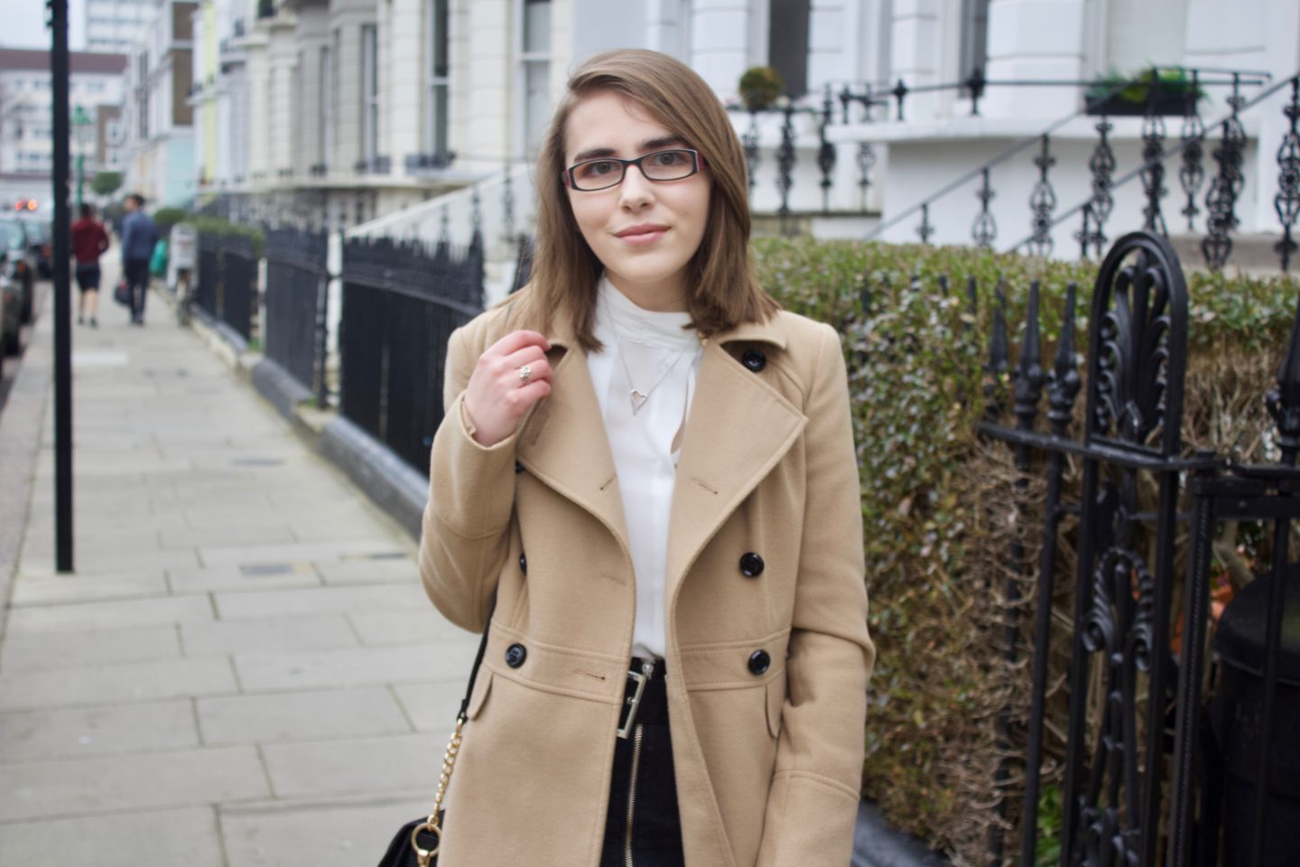 Elin wearing a camel coat and white pleated high neck blouse, she is standing in front of black railings and white houses