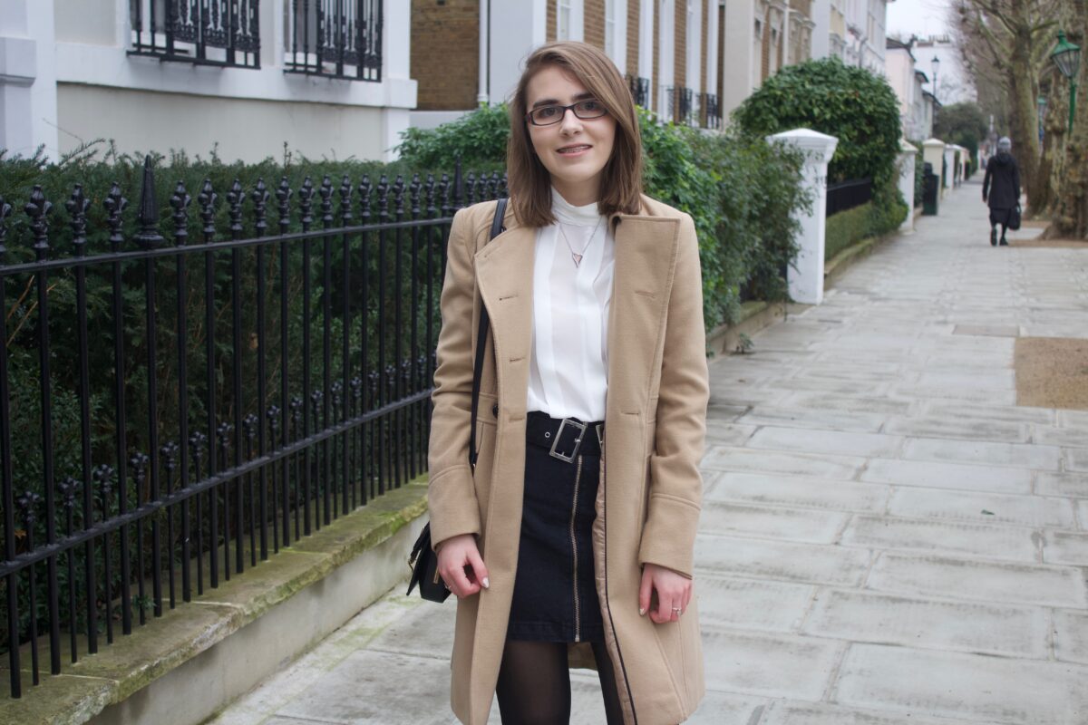 A photo of Elin wearing a camel coat, white high neck pleated blouse and a black denim skirt, she is standing on a street with pretty houses on the left hand side