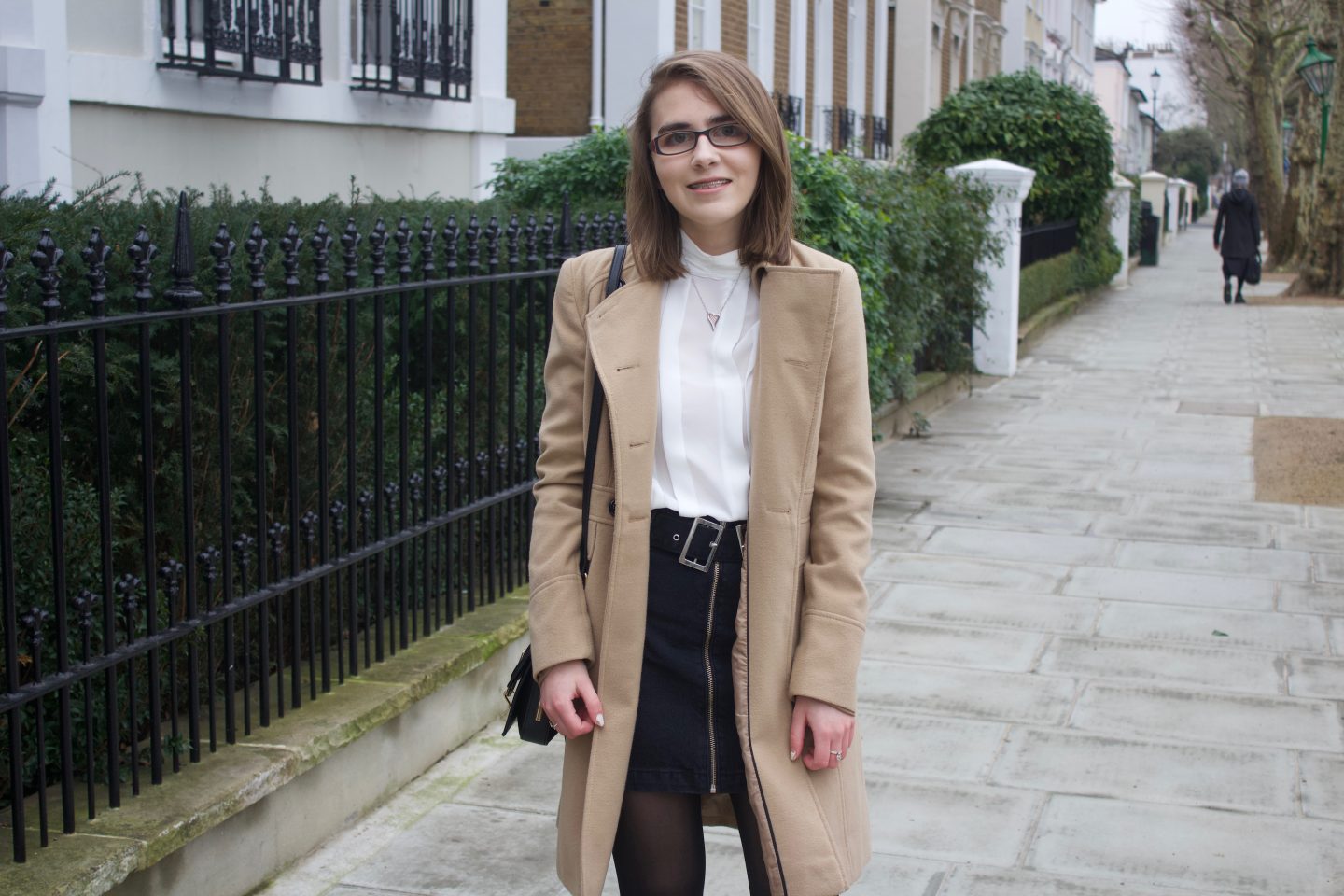 A photo of Elin wearing a camel coat, white high neck pleated blouse and a black denim skirt, she is standing on a street with pretty houses on the left hand side