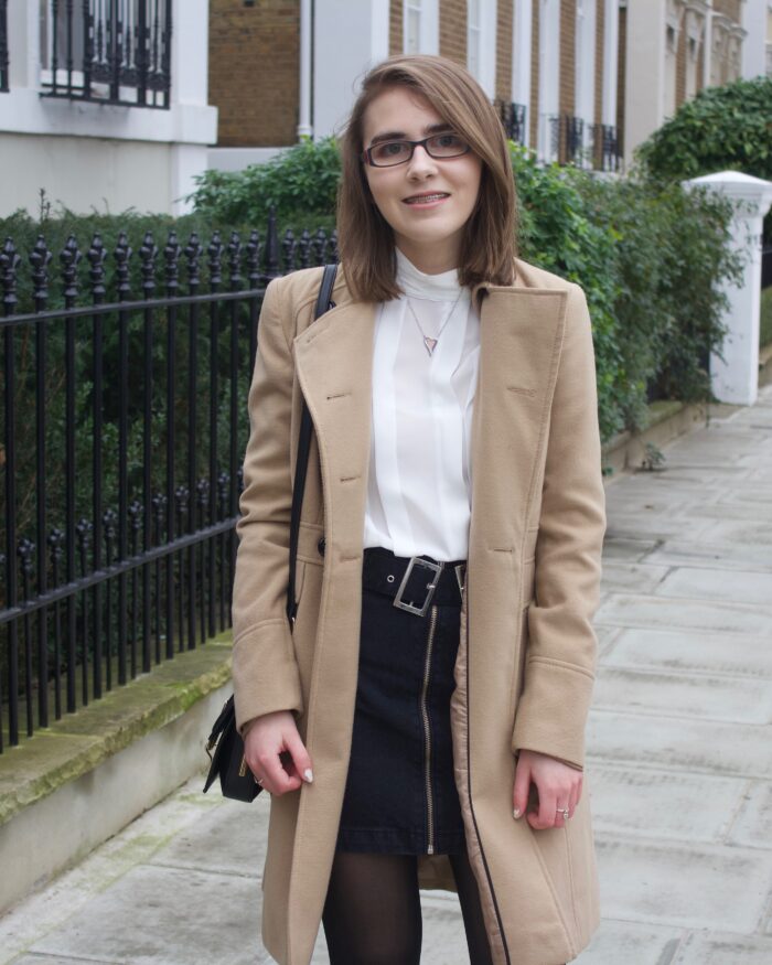 A photo of Elin wearing a camel coat, white high neck pleated blouse and a black denim skirt, she is standing on a street with pretty houses on the left hand side