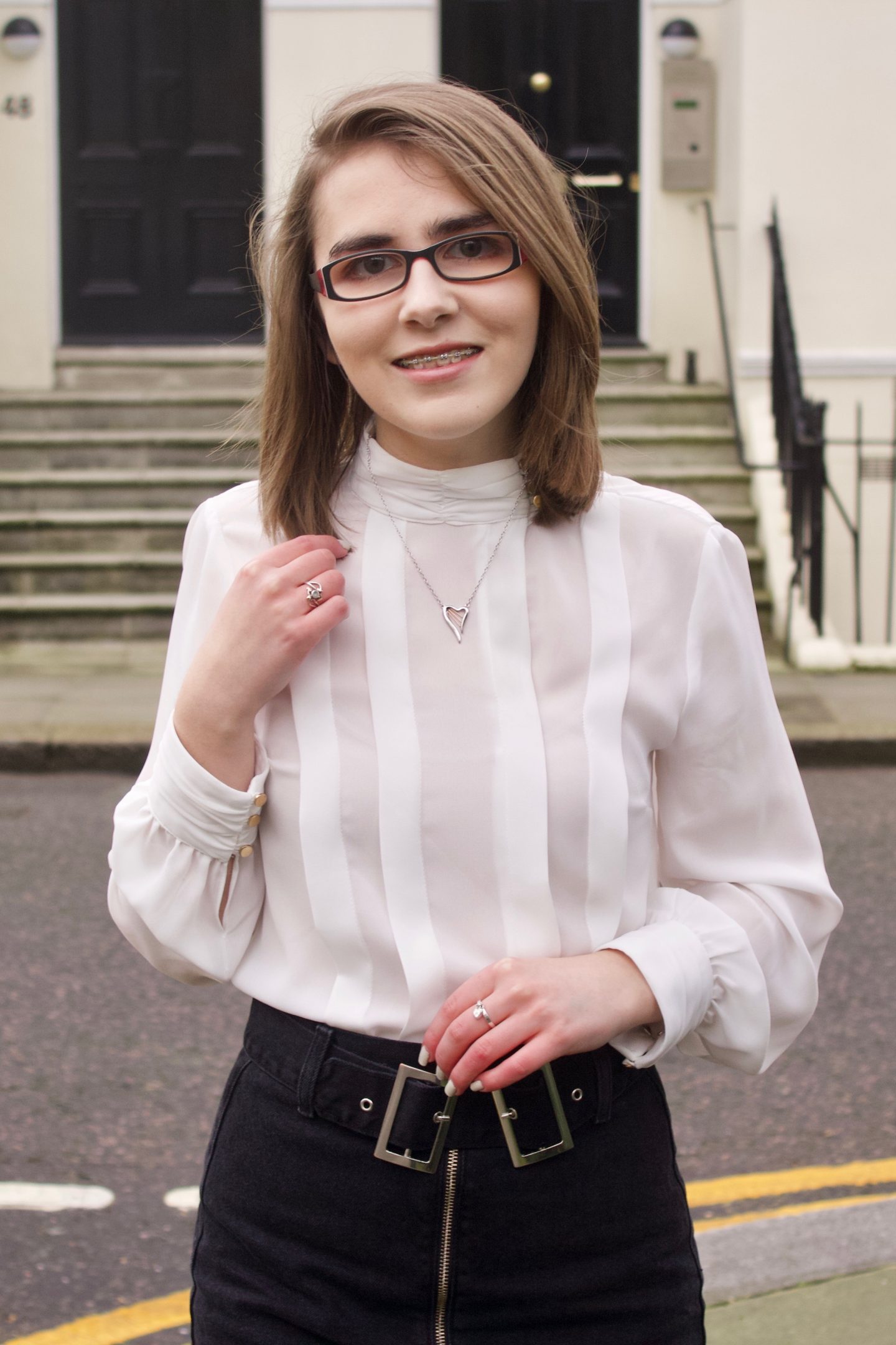 A portrait photo of Elin wearing pleated high neck blouse, a black denim zip up skirt and a harp shaped silver necklace