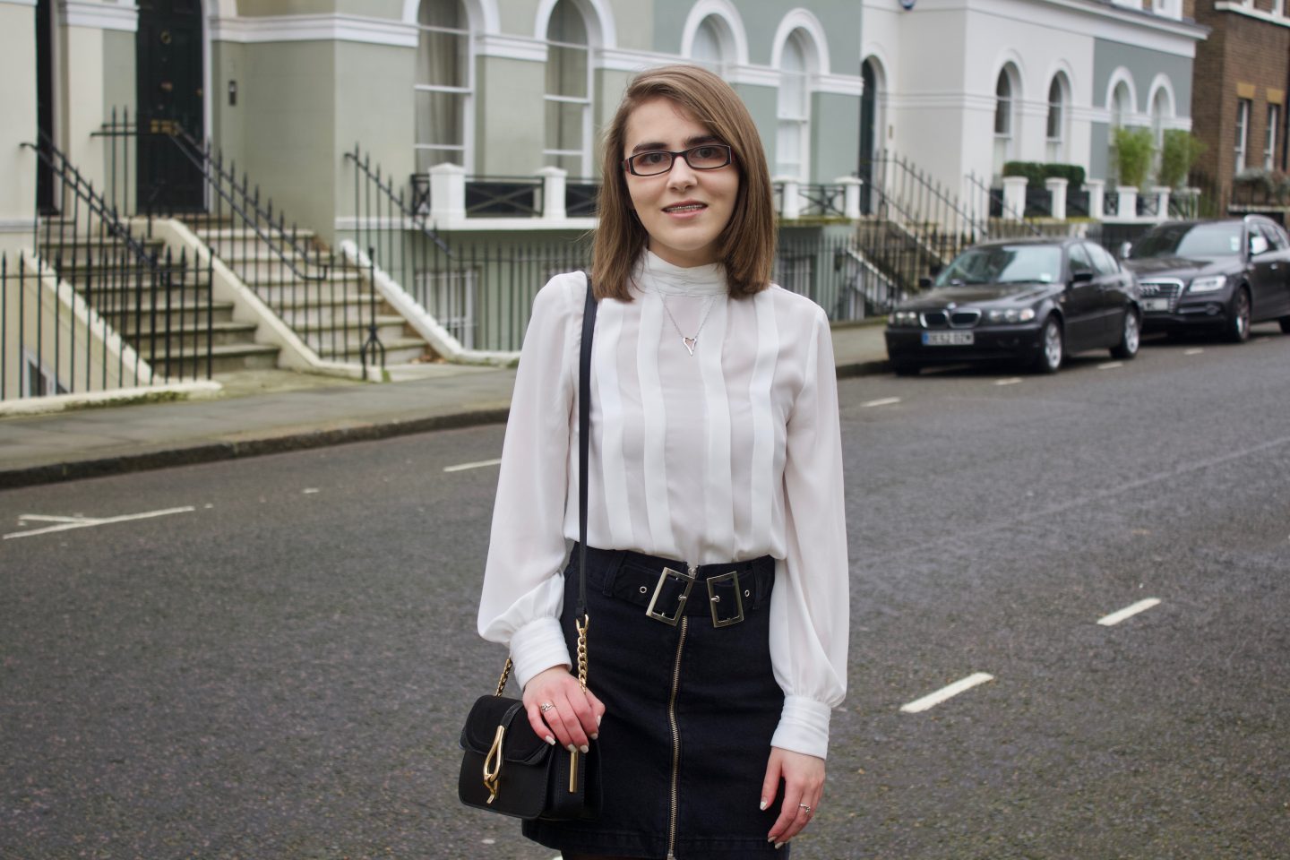 A photo of Elin wearing a black denim double buckle skirt, a white high neck pleated blouse and black cross body bag which has a gold buckle on it