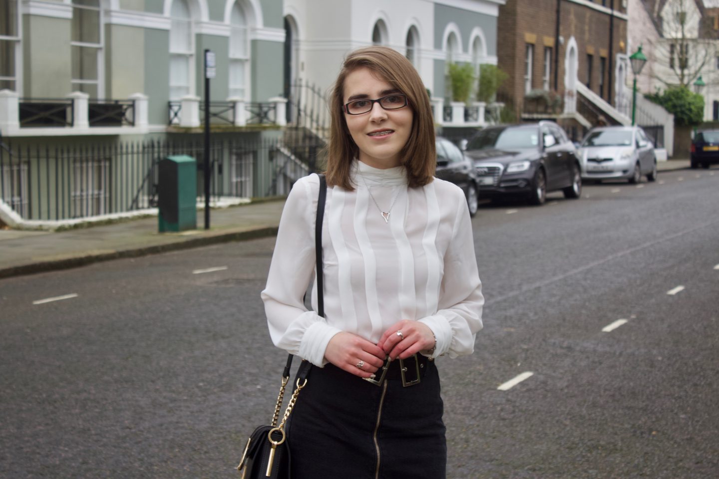 A photo of Elin wearing a black denim double buckle skirt, a white high neck pleated blouse and a black cross body bag