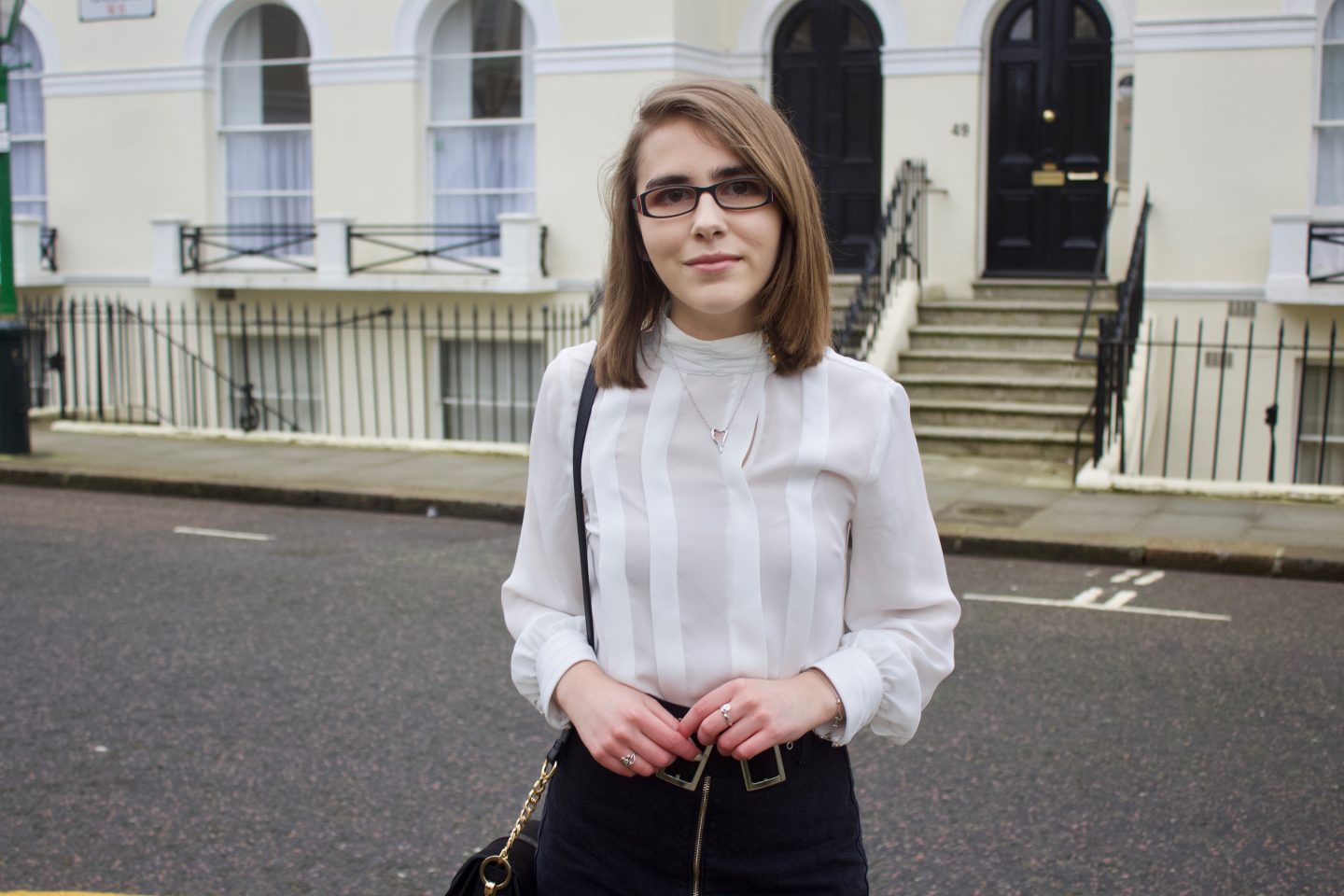 A photo of Elin wearing a white high neck pleated blouse and black denim double buckle skirt, she is standing in front of some houses which have steps leading up to them