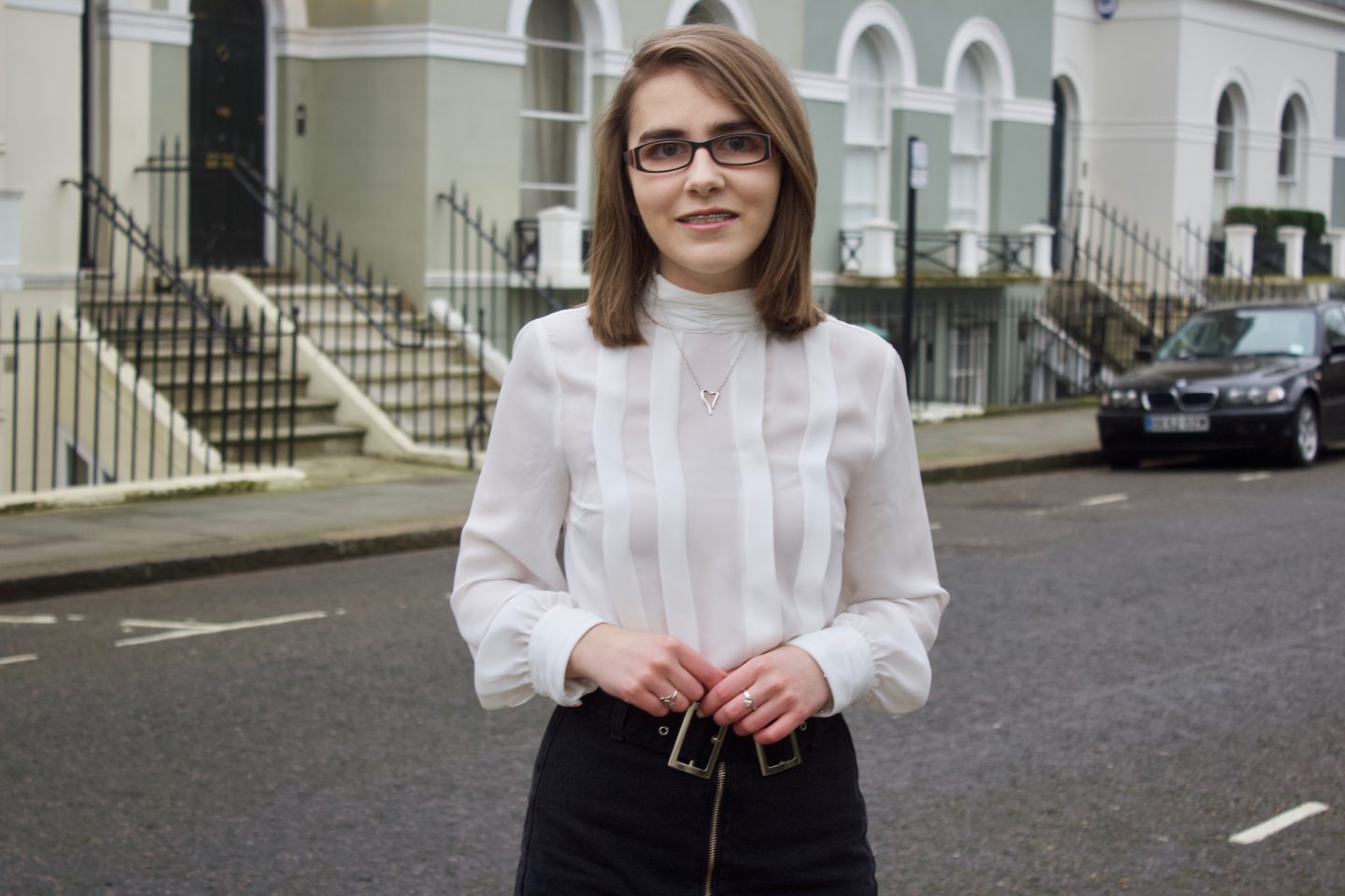 A photo of Elin standing in front of some houses with steps leading up to them in London, she is wearing a white high neck pleated blouse and a zip up denim belt which has a double buckle belt