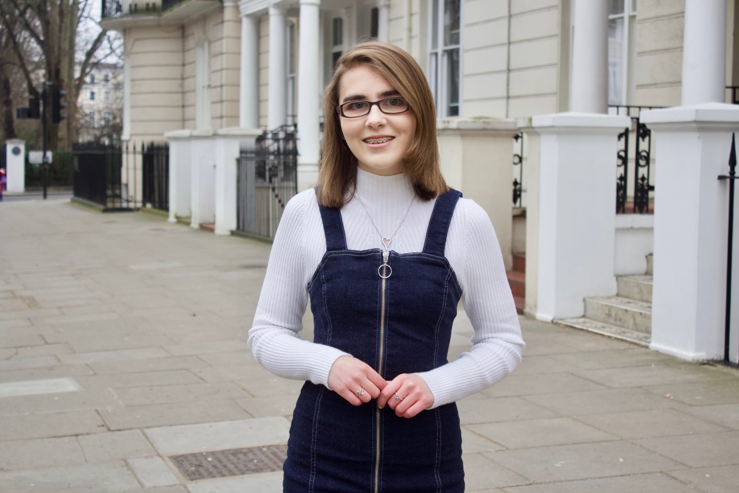 A photo of Elin smiling at the camera, she is wearing a black pinafore which has a zip down the front, a white high neck top and a harp shaped necklace