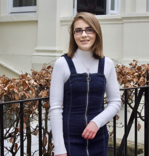 A photo of Elin standing in front of black railings which has pink flowers along the top