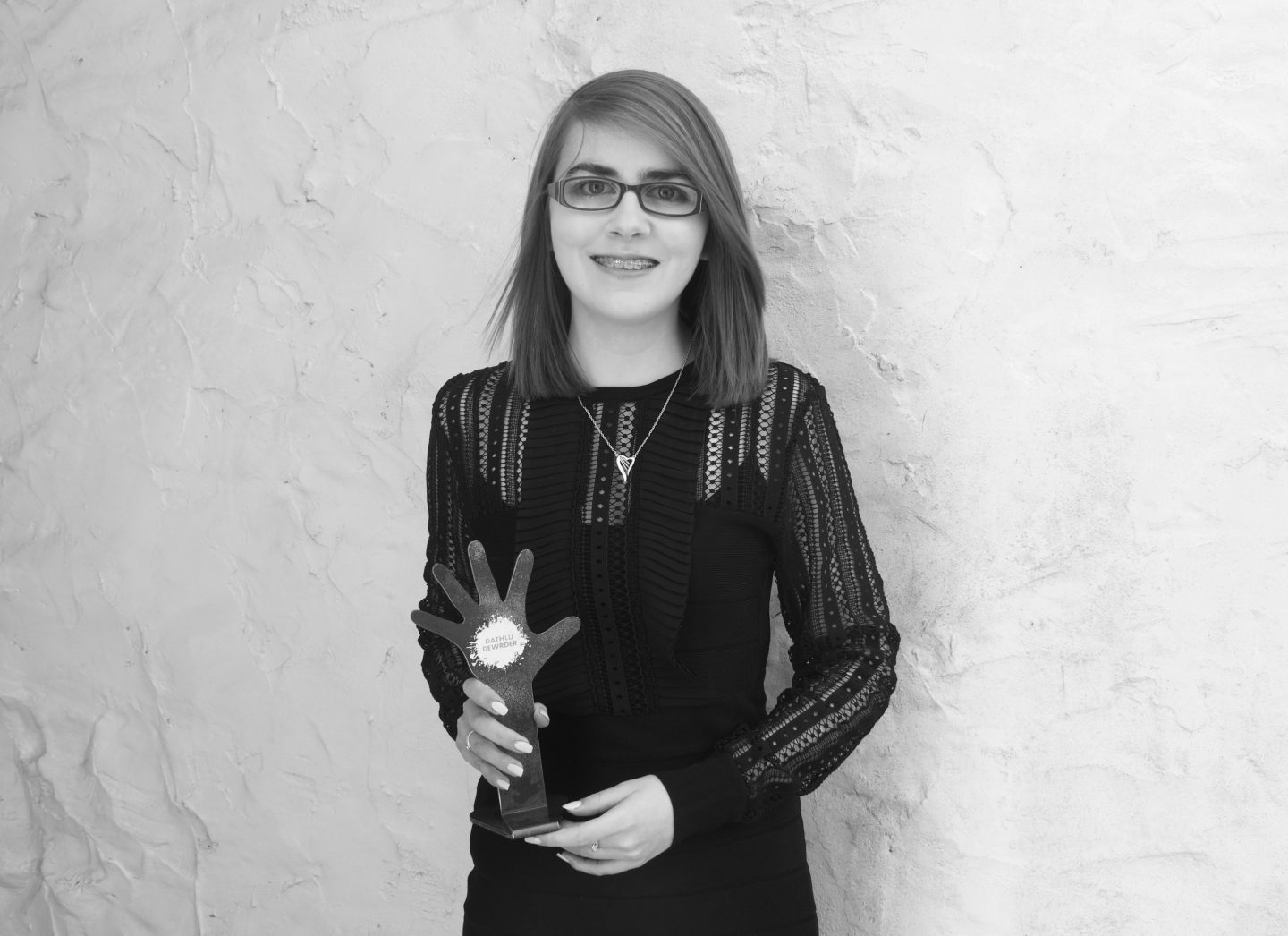 A photo of Elin holding her 'Dathlu Dewrder' award which is in a shape of a hand being raised. She wears a black French Connection dress which has lace along the sleeves and neckline.