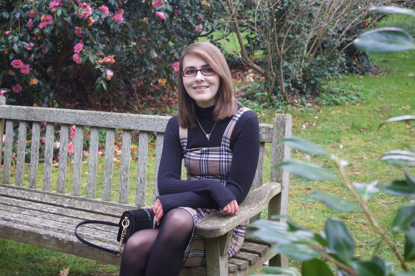 A photo of Elin sitting on a bench, she is wearing a beige check pinafore over a black high neck top