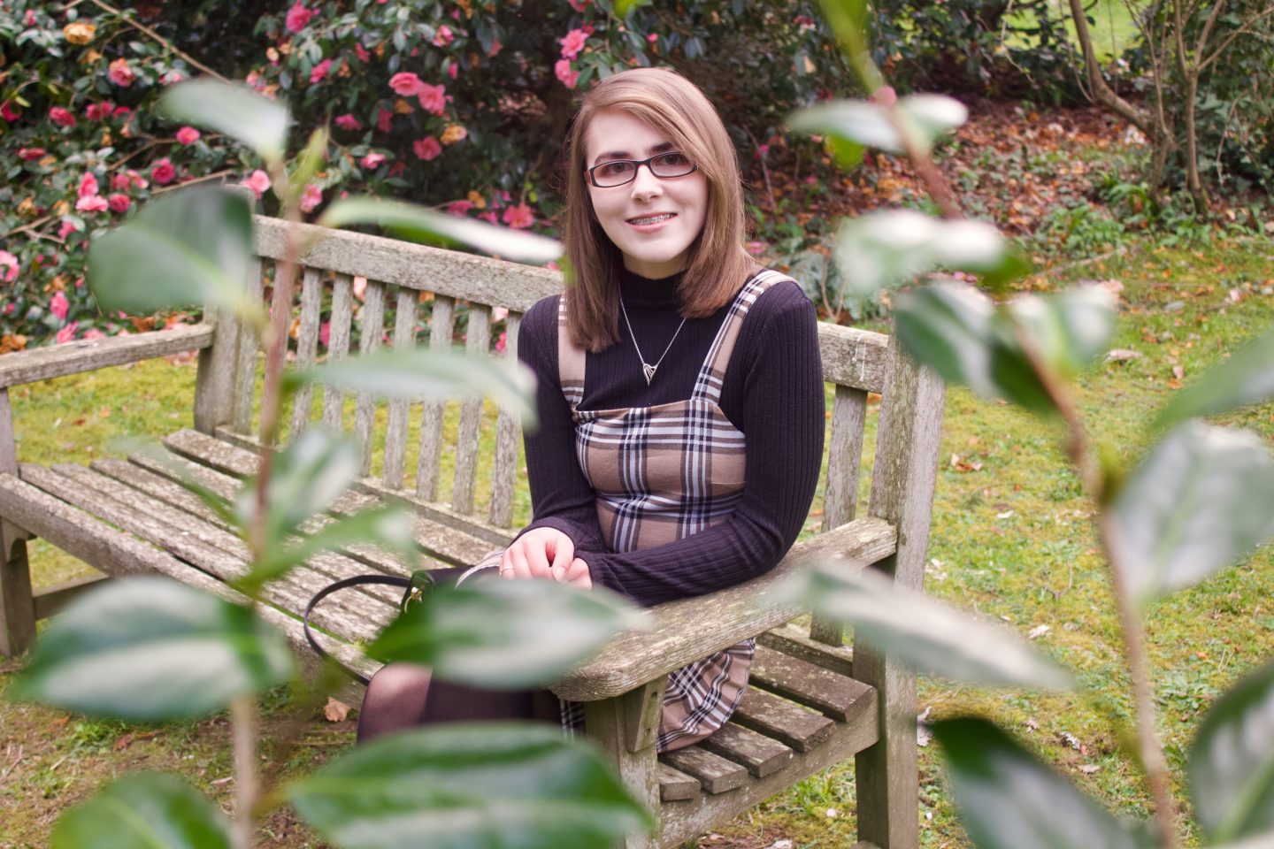 A photo of Elin sitting on a bench with trees and flowers in the background and some leaves in the foreground which are out of fovus